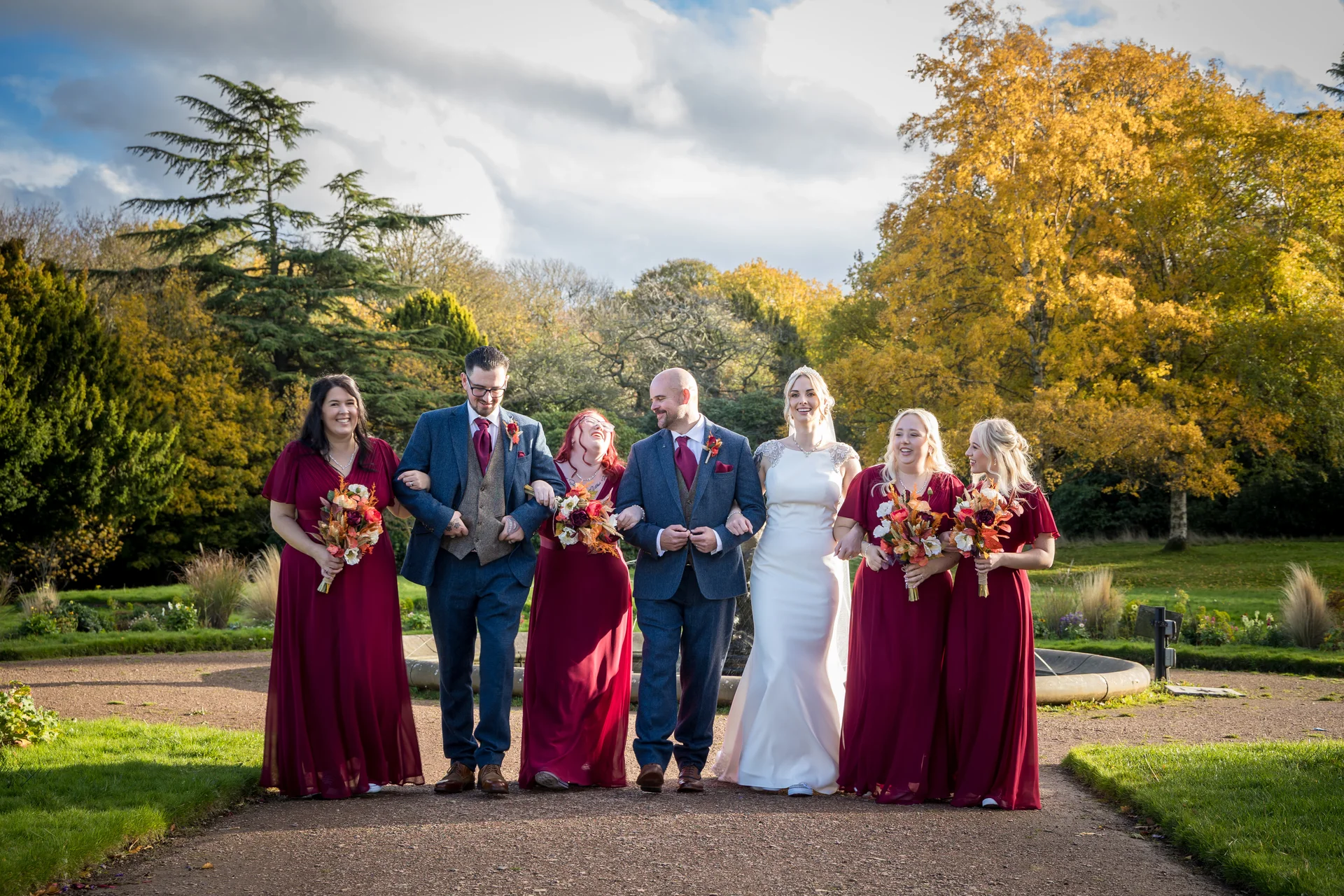 The wedding party strolls along a garden path surrounded by vibrant autumn foliage, with the bride and groom laughing amongst their bridesmaids and groomsmen.