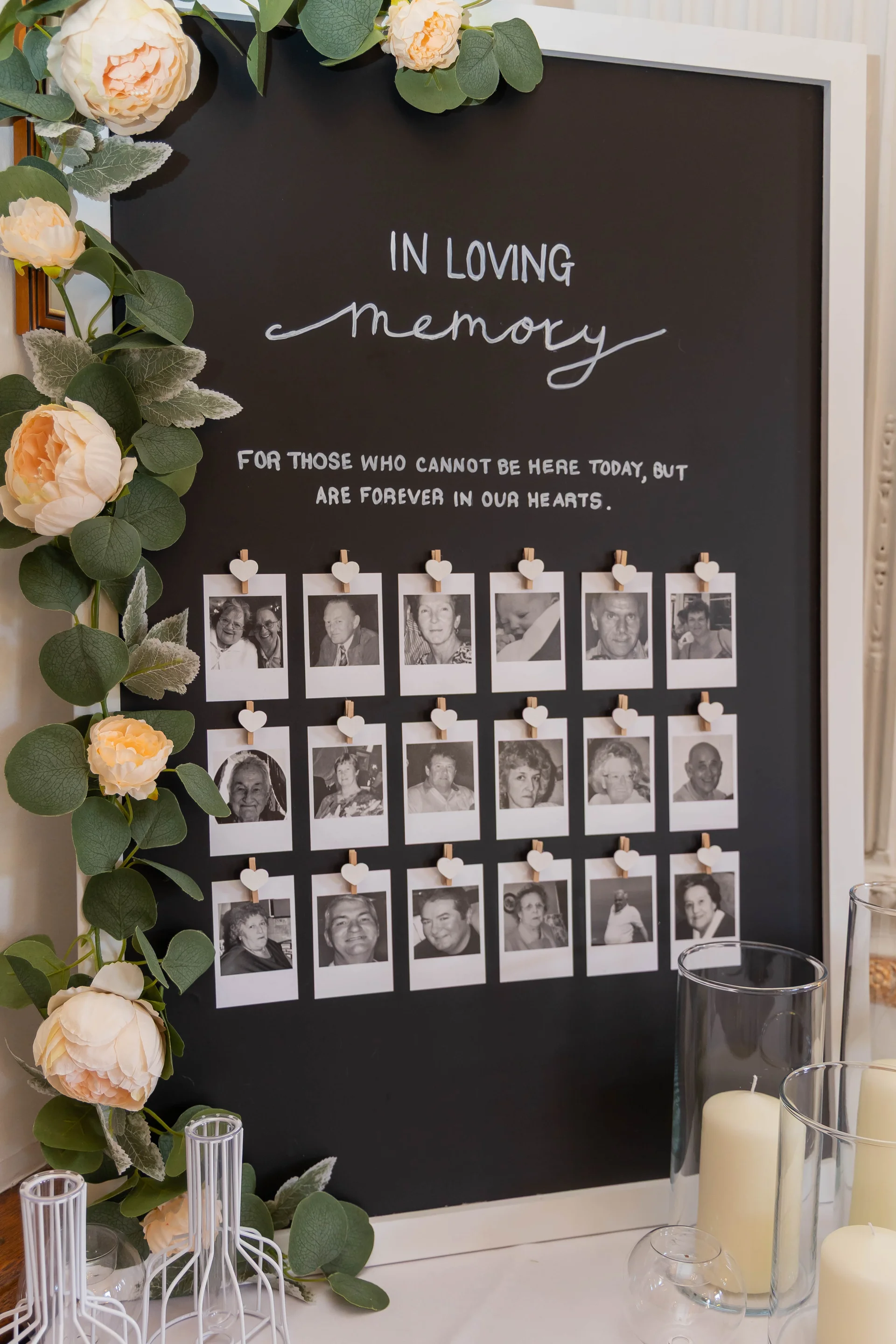 A memory table display featuring a black board with handwritten lettering and black-and-white photos attached with tiny wooden heart pegs, adorned with blush roses and eucalyptus.