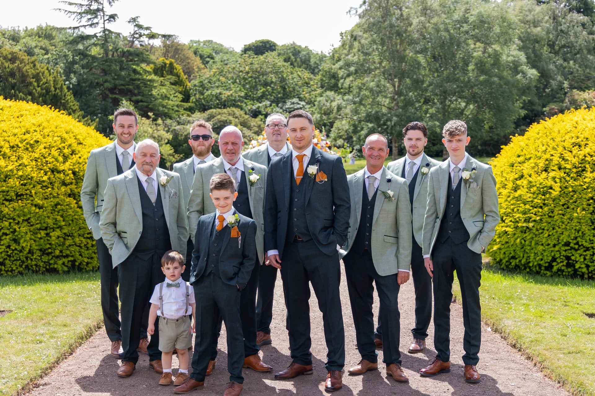 A groom and his large group of groomsmen and page boys pose outdoors in matching green suits and orange ties on a sunny day.