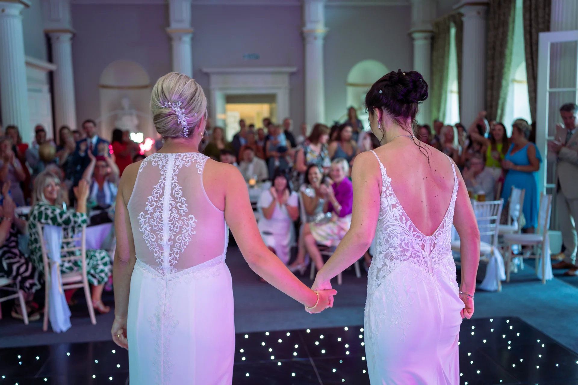 Two brides in white lace wedding dresses hold hands facing their cheering guests on a lighted dance floor in a grand ballroom.