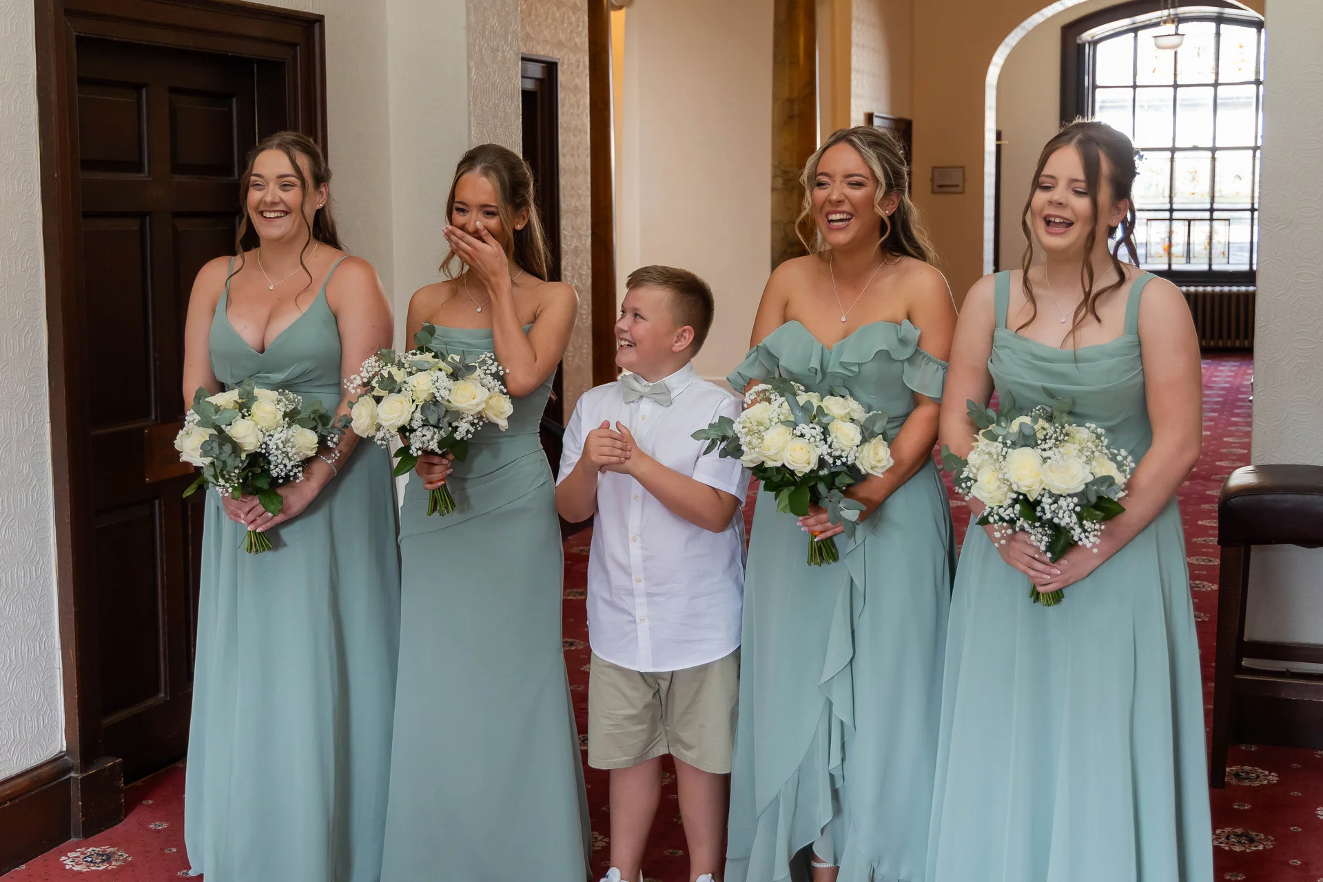 Four bridesmaids in sage green dresses and a young page boy stand together in a hotel hallway, laughing joyfully while holding bouquets of white roses.