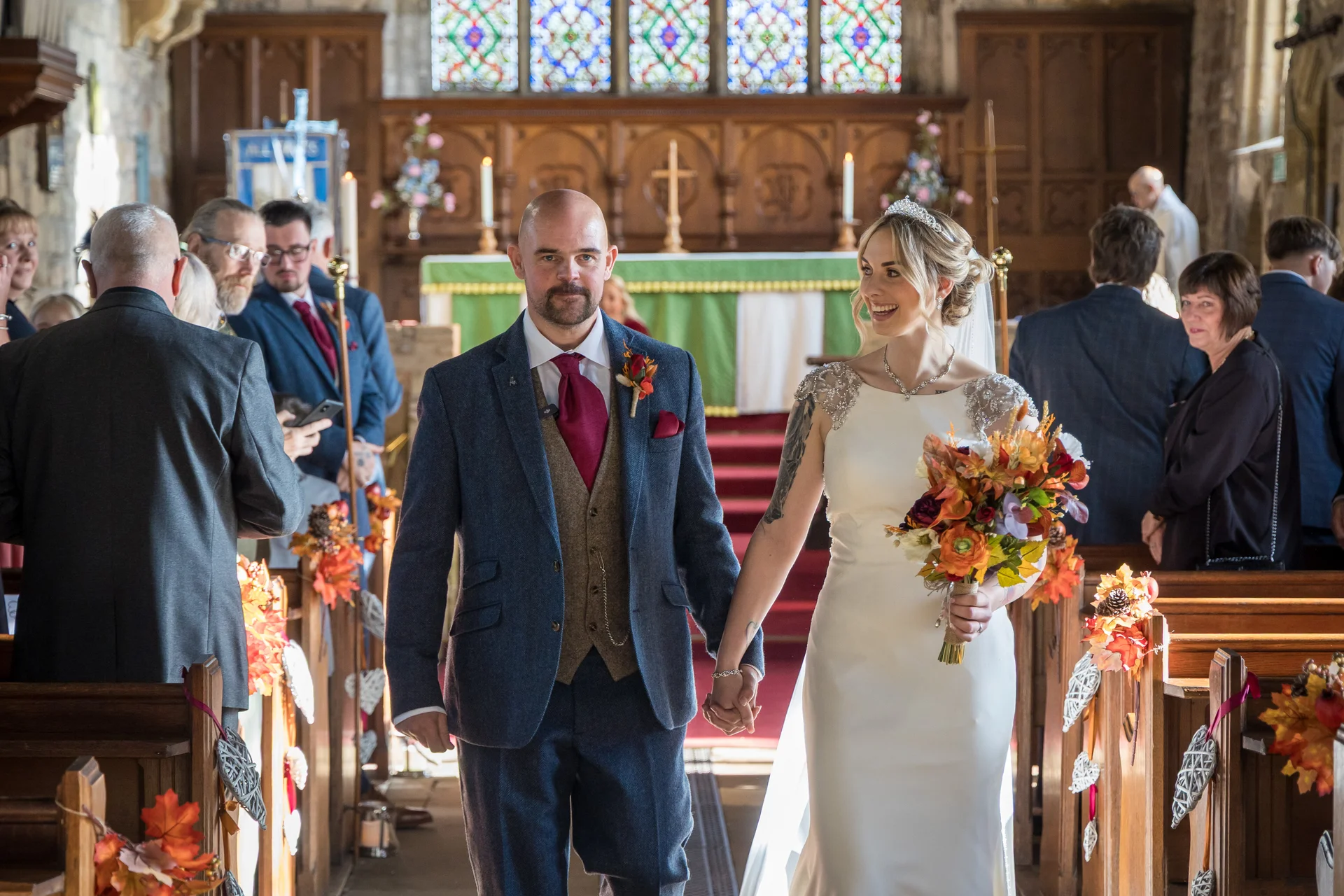 A smiling newlywed couple walks hand in hand down the aisle of a historic church decorated with autumn foliage after their ceremony.