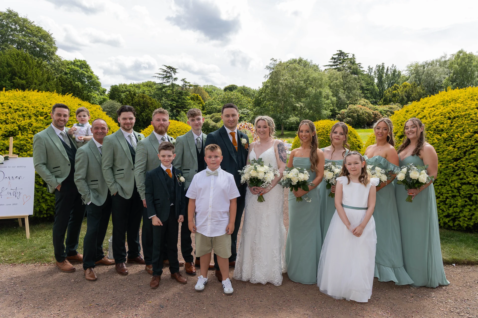 A large wedding party including the bride and groom, groomsmen in sage green suits, bridesmaids in matching sage dresses, and young attendants posing outdoors in a lush garden setting.