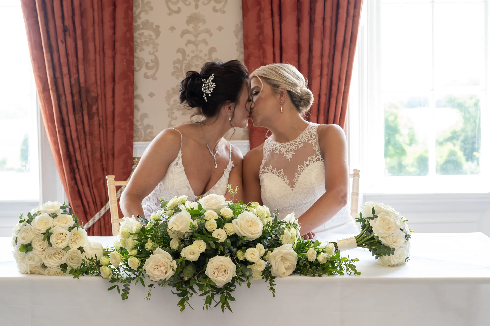 Two brides share a tender kiss seated at a table adorned with lush white roses and eucalyptus foliage inside an elegant venue with draped red curtains.