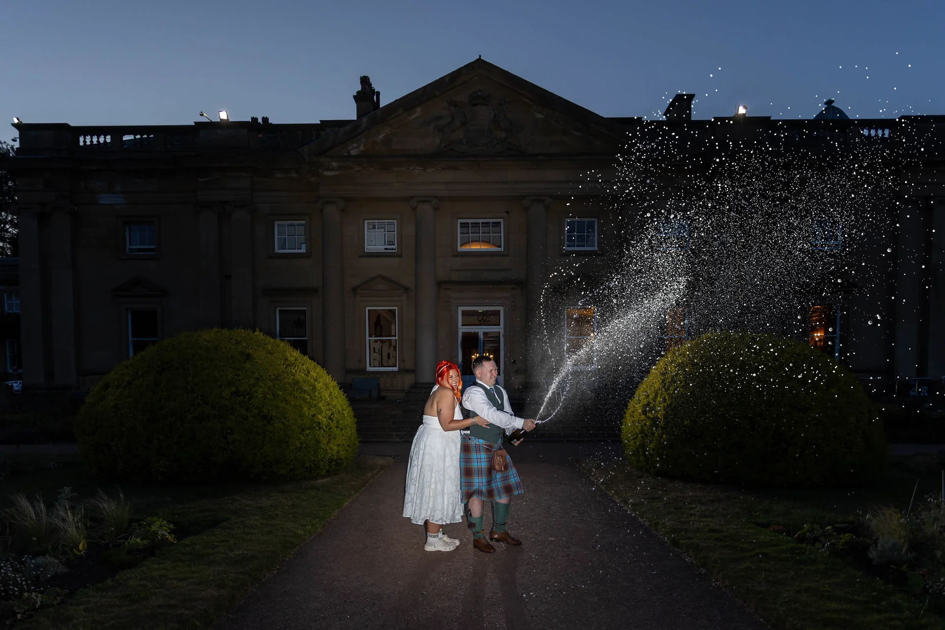A newlywed couple excitedly spraying champagne outside a grand country house during blue hour, with the groom wearing a traditional tartan kilt and the bride in a white dress with pink hair.