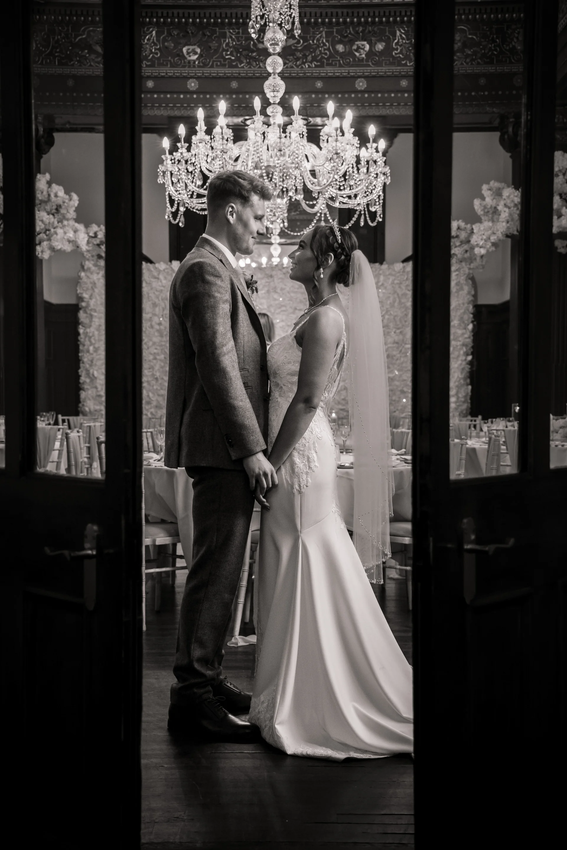 A newlywed couple holds hands and gazes at each other inside a grand room illuminated by a crystal chandelier, viewed through open double doors.