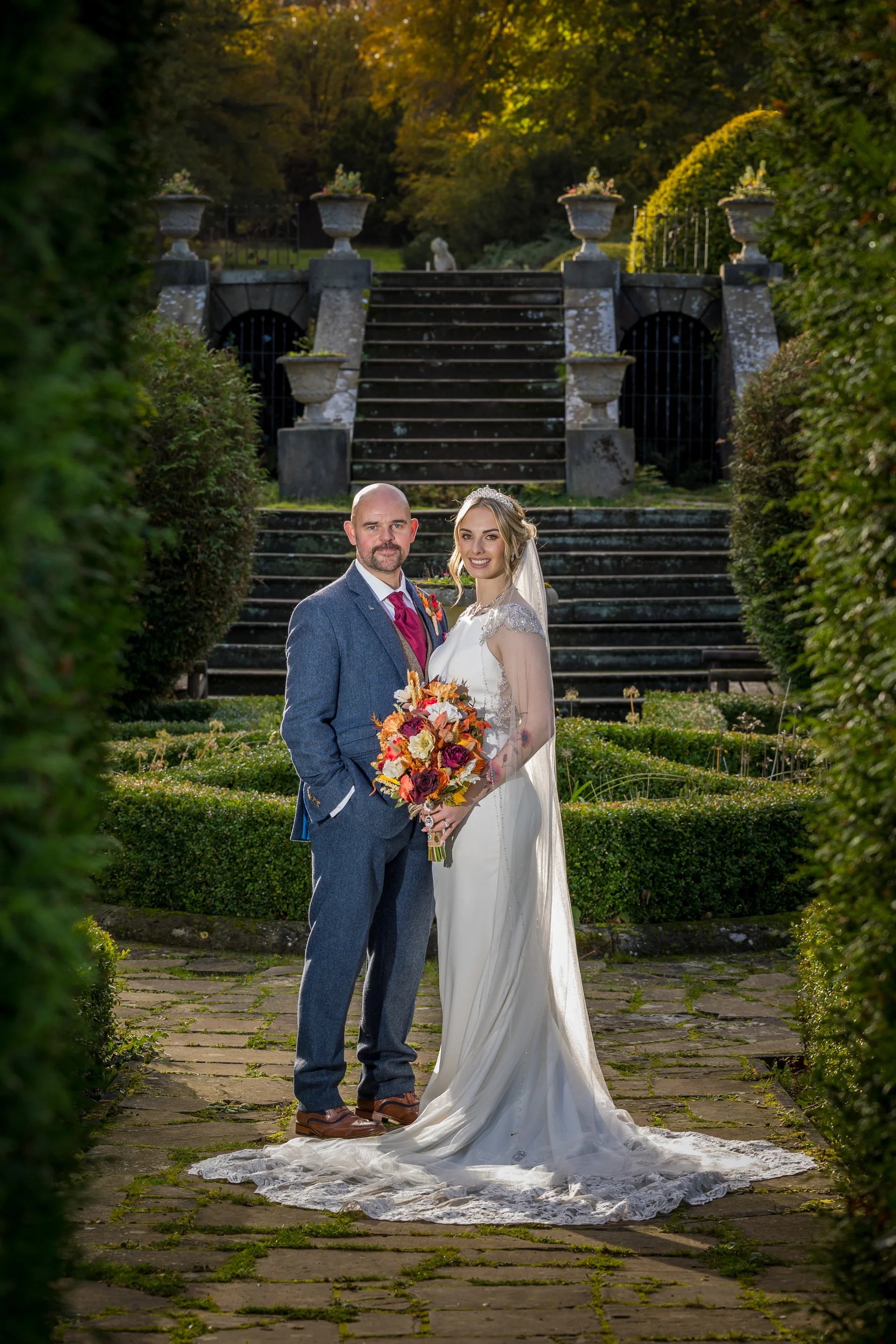 A newlywed couple poses together on a stone pathway framed by garden topiary, with the bride holding an autumnal bouquet and wearing an embellished gown and veil.