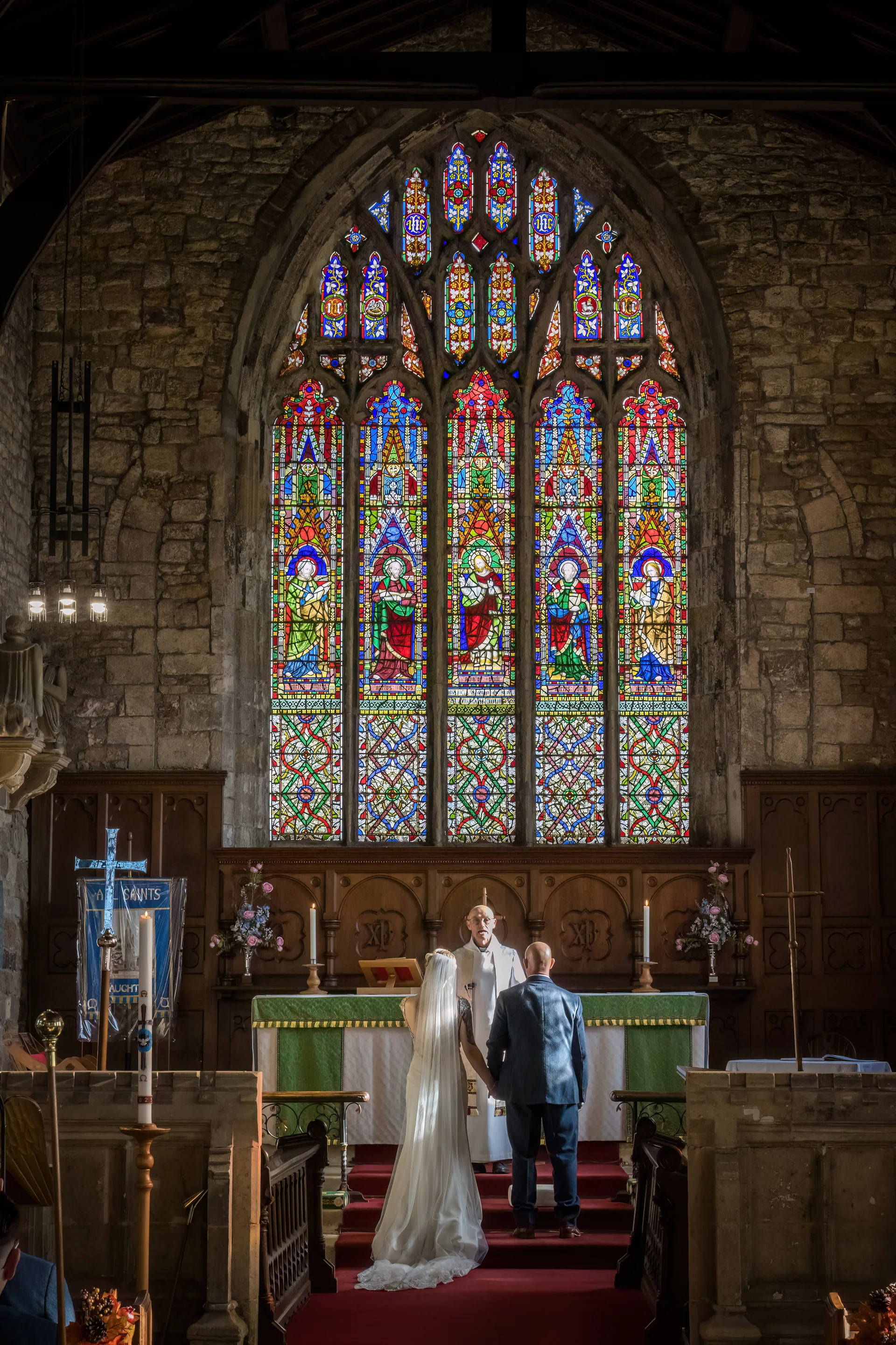 A bride and groom stand holding hands at the altar during their church wedding ceremony, framed by a magnificent stained-glass window and historic stone architecture.