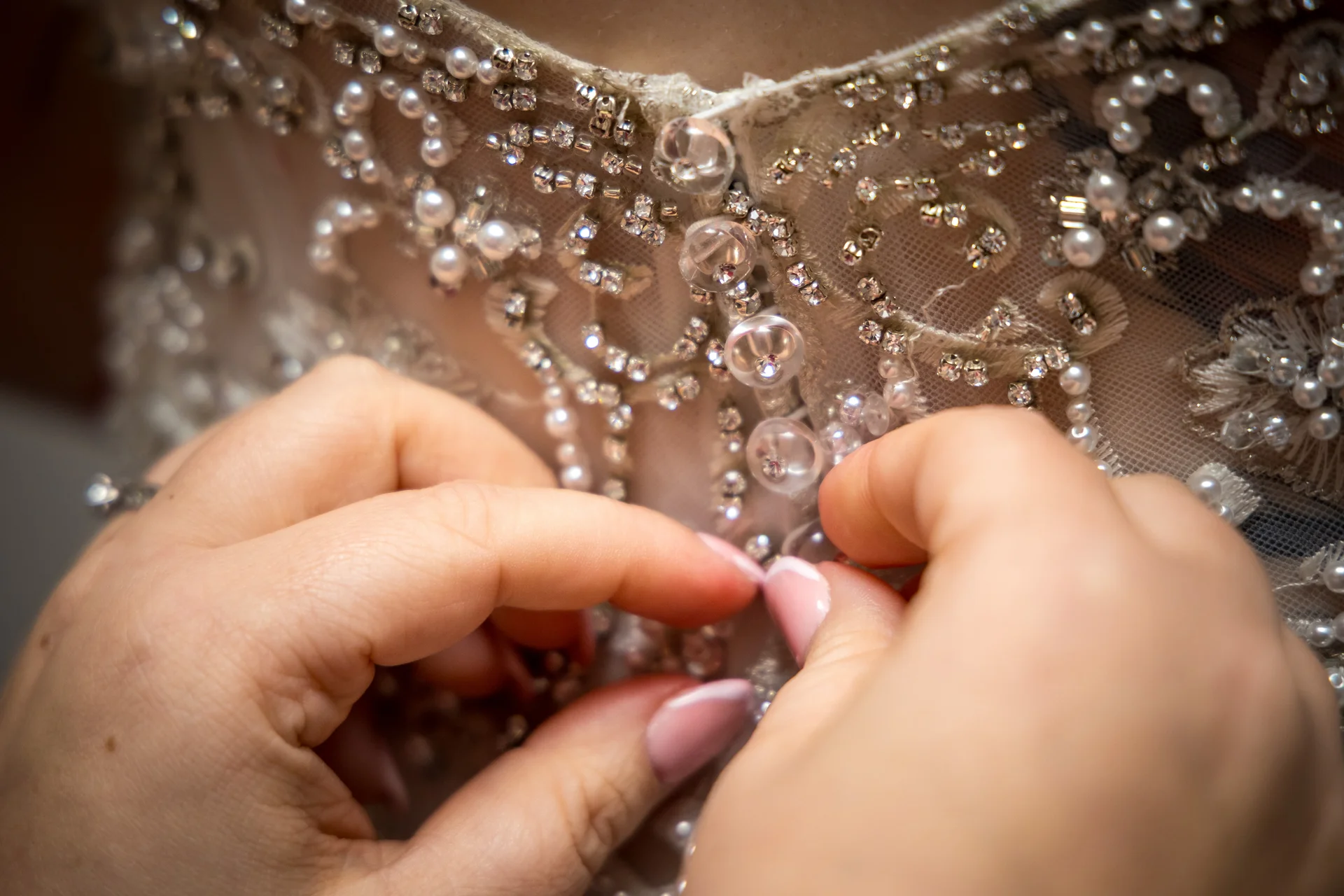 Close-up of hands carefully fastening the delicate crystal and pearl-embellished buttons on the back of a bridal gown during preparations.