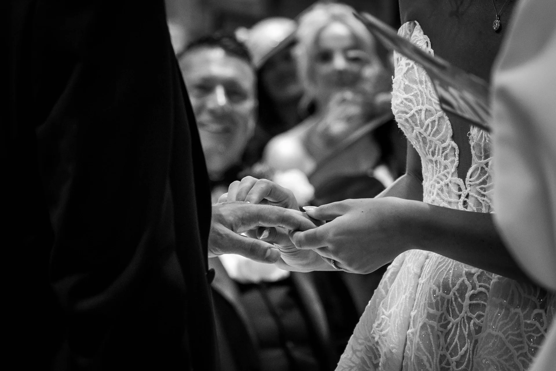 An intimate black and white shot captures the bride and groom exchanging rings at the altar, framed by smiling wedding guests in the background.