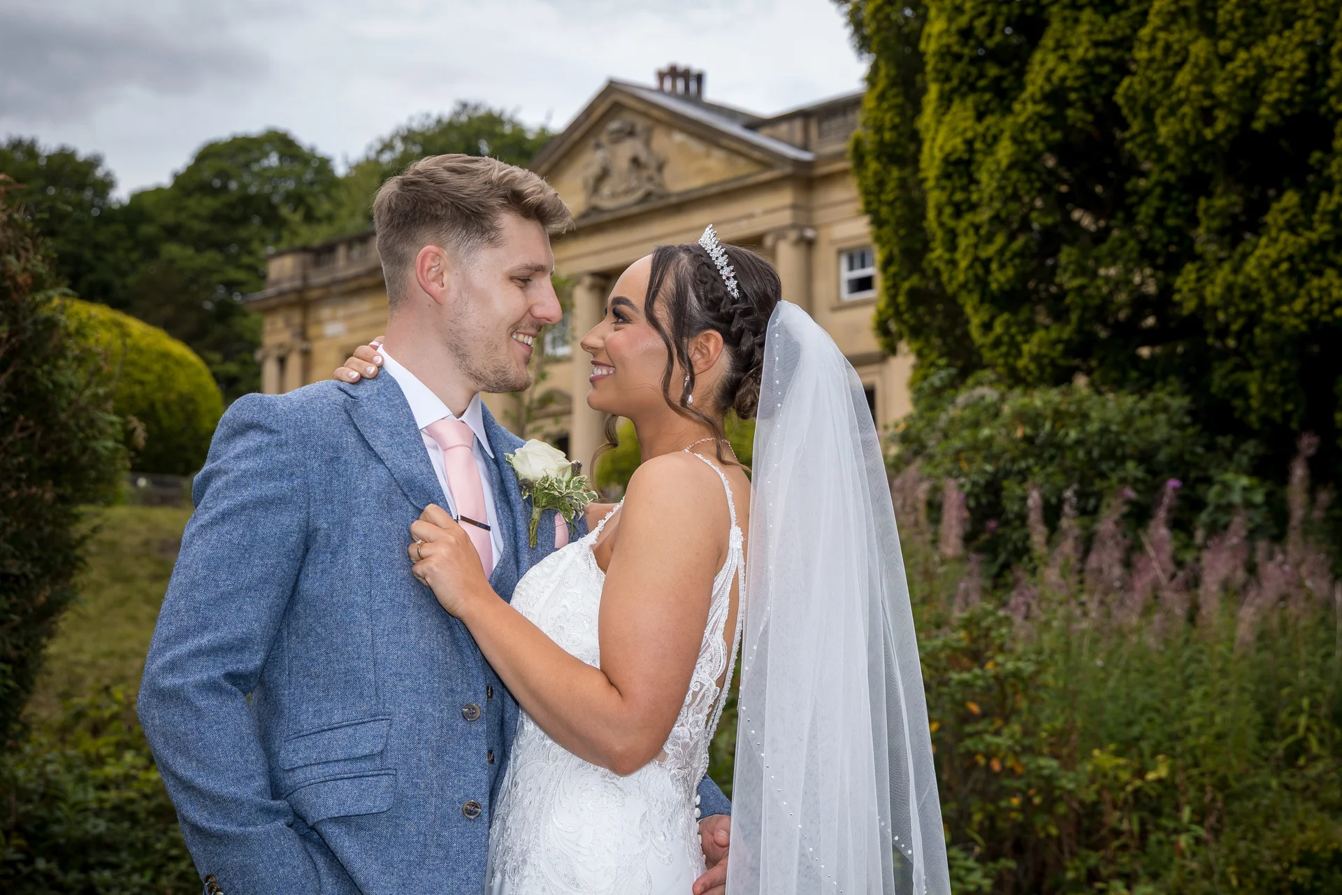 A newlywed couple smiles at each other outside a grand stone manor house, with the groom wearing a blue tweed suit and pink tie, and the bride in a lace wedding dress and veil.