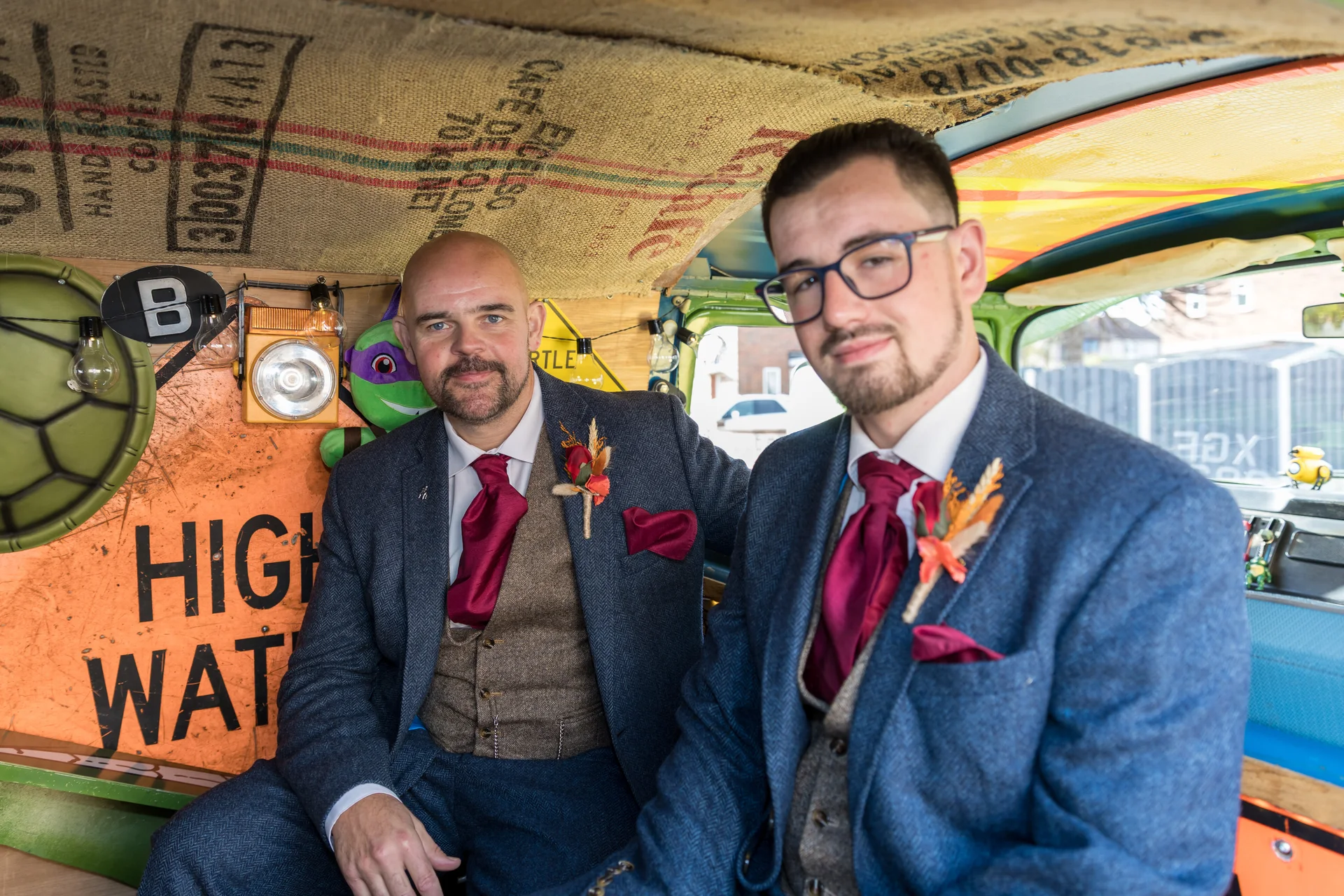 Two grooms in matching blue tweed suits and burgundy ties sit inside a uniquely decorated campervan with hessian lining and rustic signage.