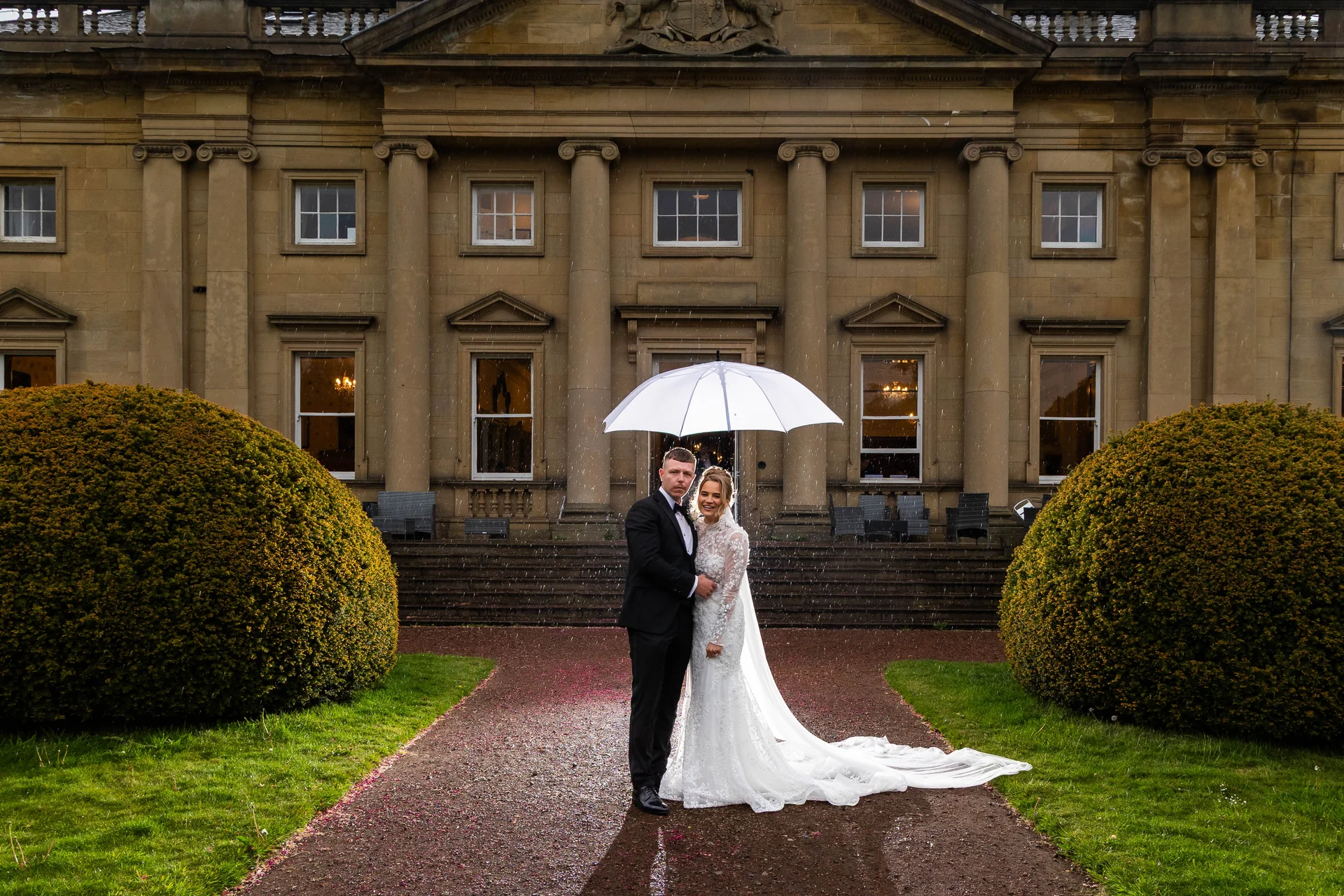 A bride and groom pose under a clear umbrella on a rainy day in front of a grand country house with manicured topiary bushes.