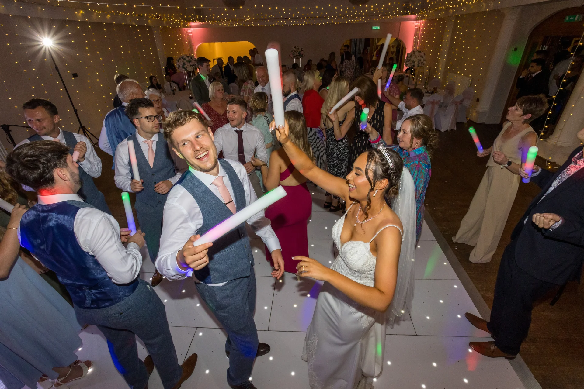 The bride and groom joyfully wave glowing foam batons while dancing with guests on a illuminated white dance floor under a canopy of fairy lights.