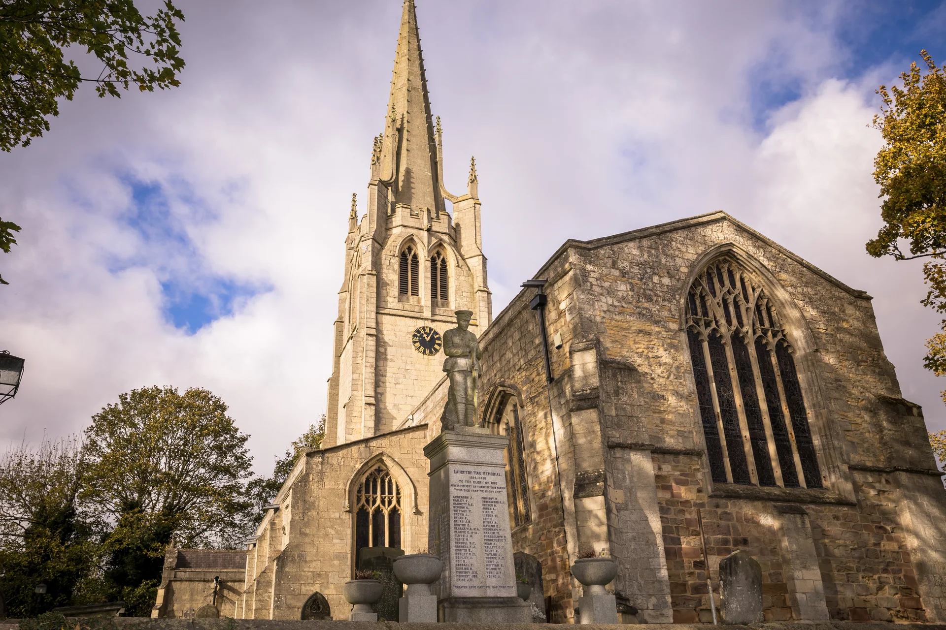 A historic stone church with a tall spire and ornate stained glass windows stands against a dramatic sky with golden autumnal trees.