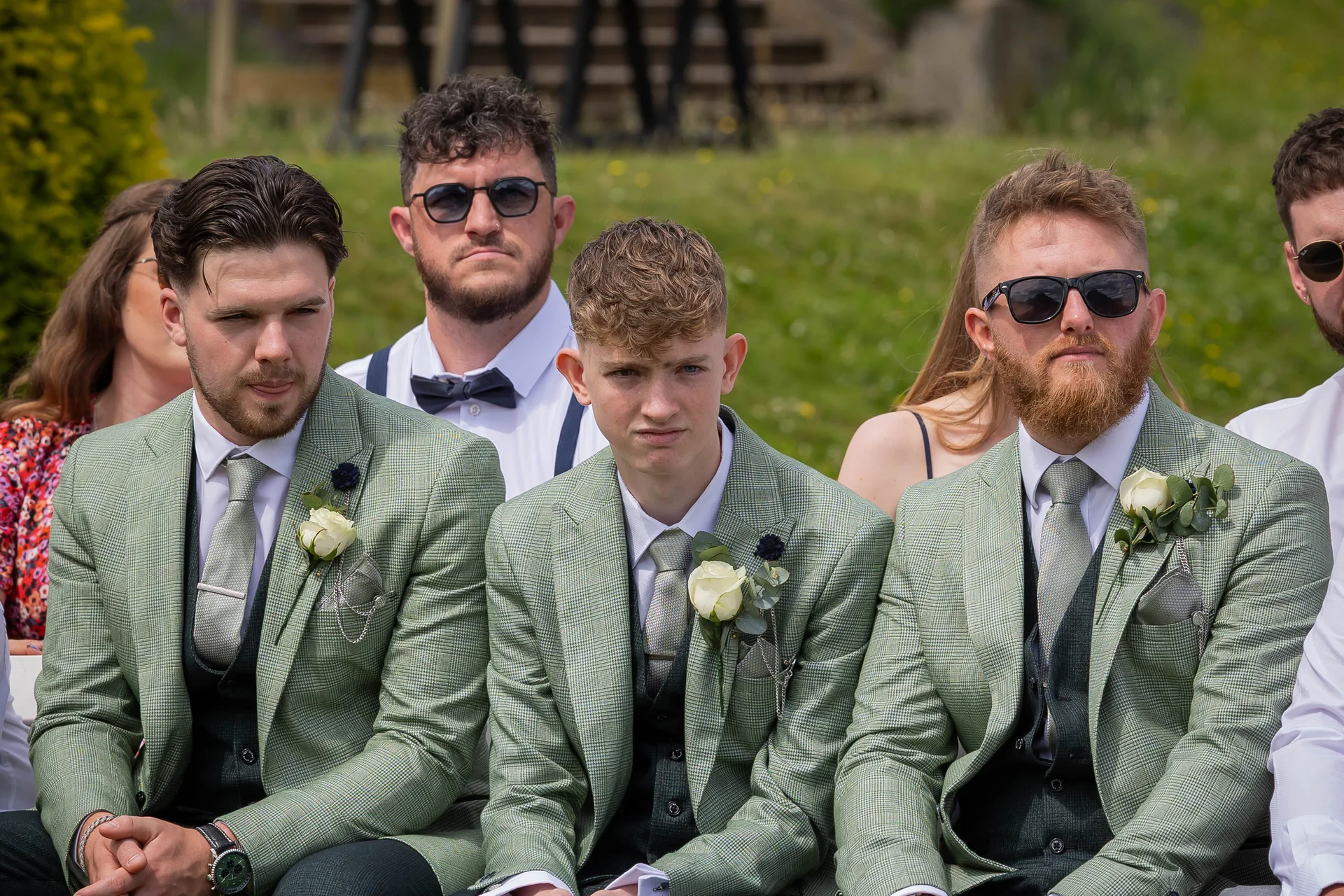 A group of groomsmen wearing matching patterned green suits, waistcoats, and white rose buttonholes seated outdoors at a wedding.