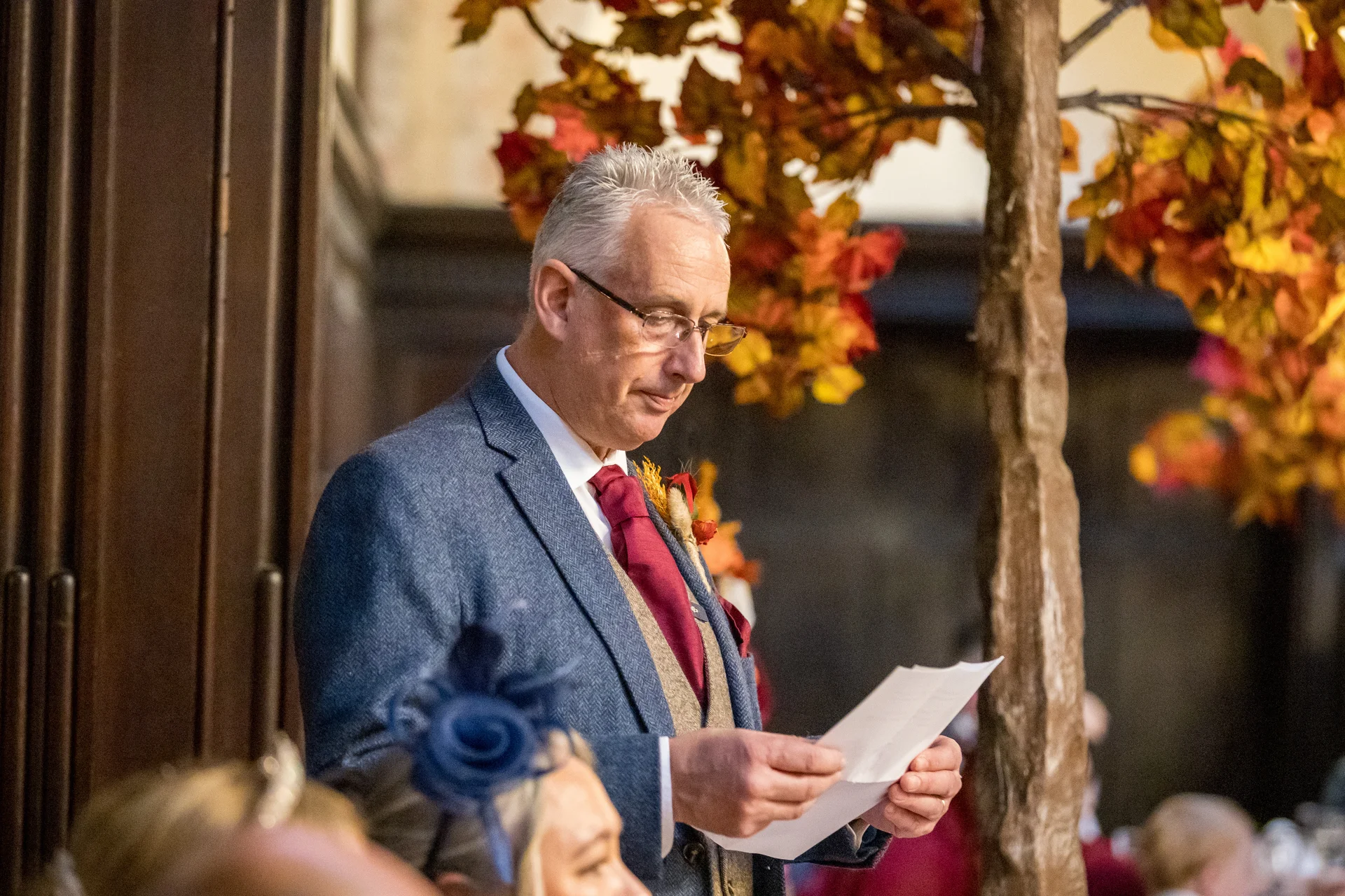A groom wearing a blue tweed suit and red tie reads a speech during the wedding reception next to an autumnal tree decoration.