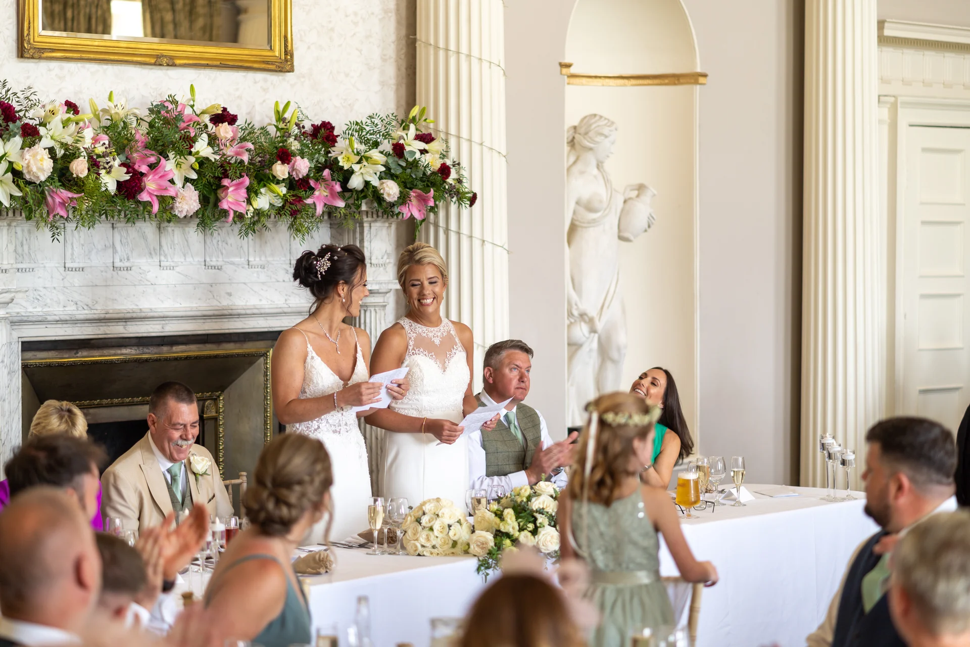 Two smiling brides deliver a speech at the top table in an elegant country house wedding reception room decorated with lush mantelpiece florals.