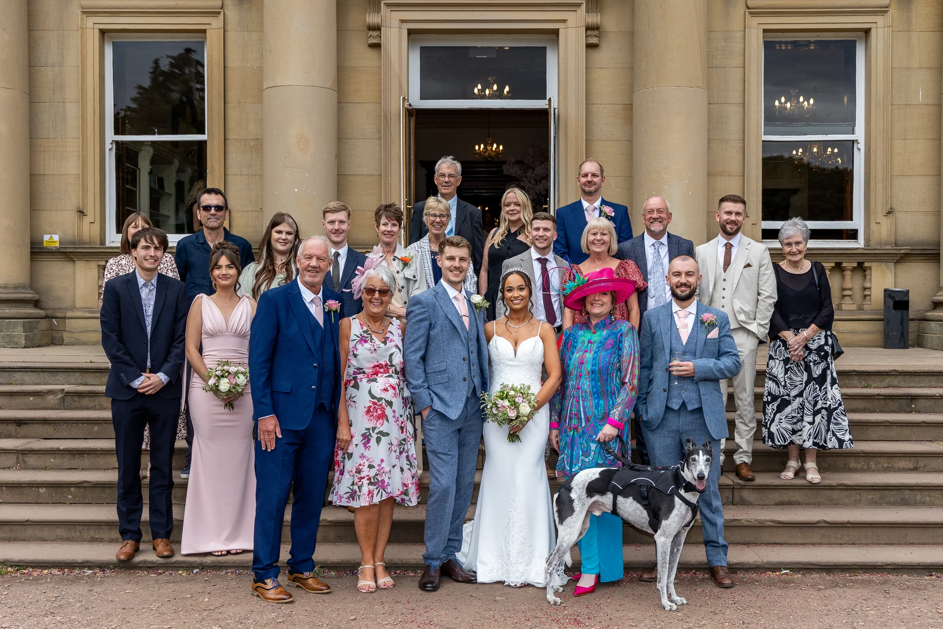 The newlywed couple stands proudly with family, wedding party, and a pet greyhound on the grand stone steps of a stately country house venue.