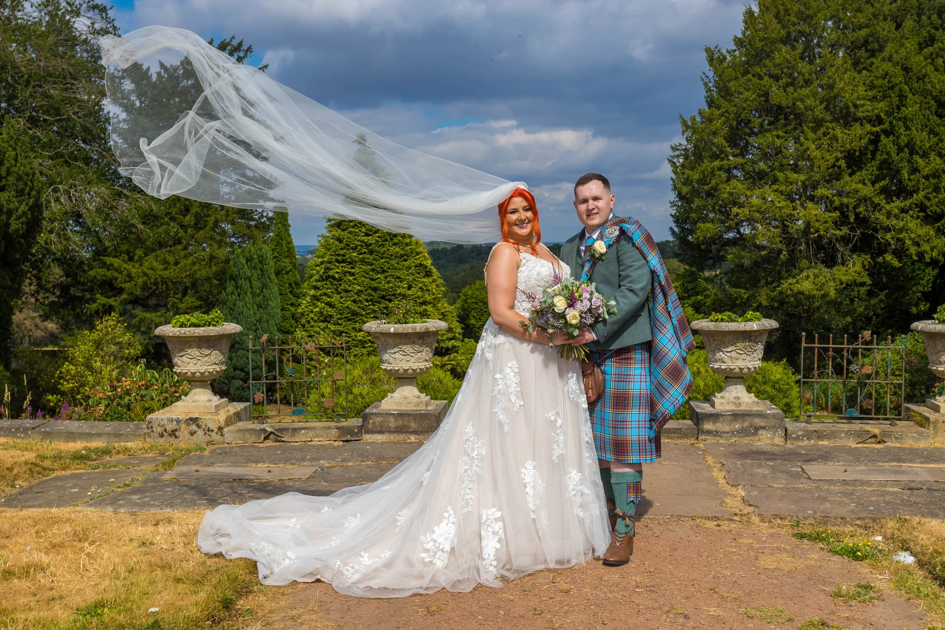 A bride with vibrant orange hair and a dramatic veil caught in the wind poses outdoors with her groom in a Scottish kilt against a dramatic cloudy sky and garden backdrop.