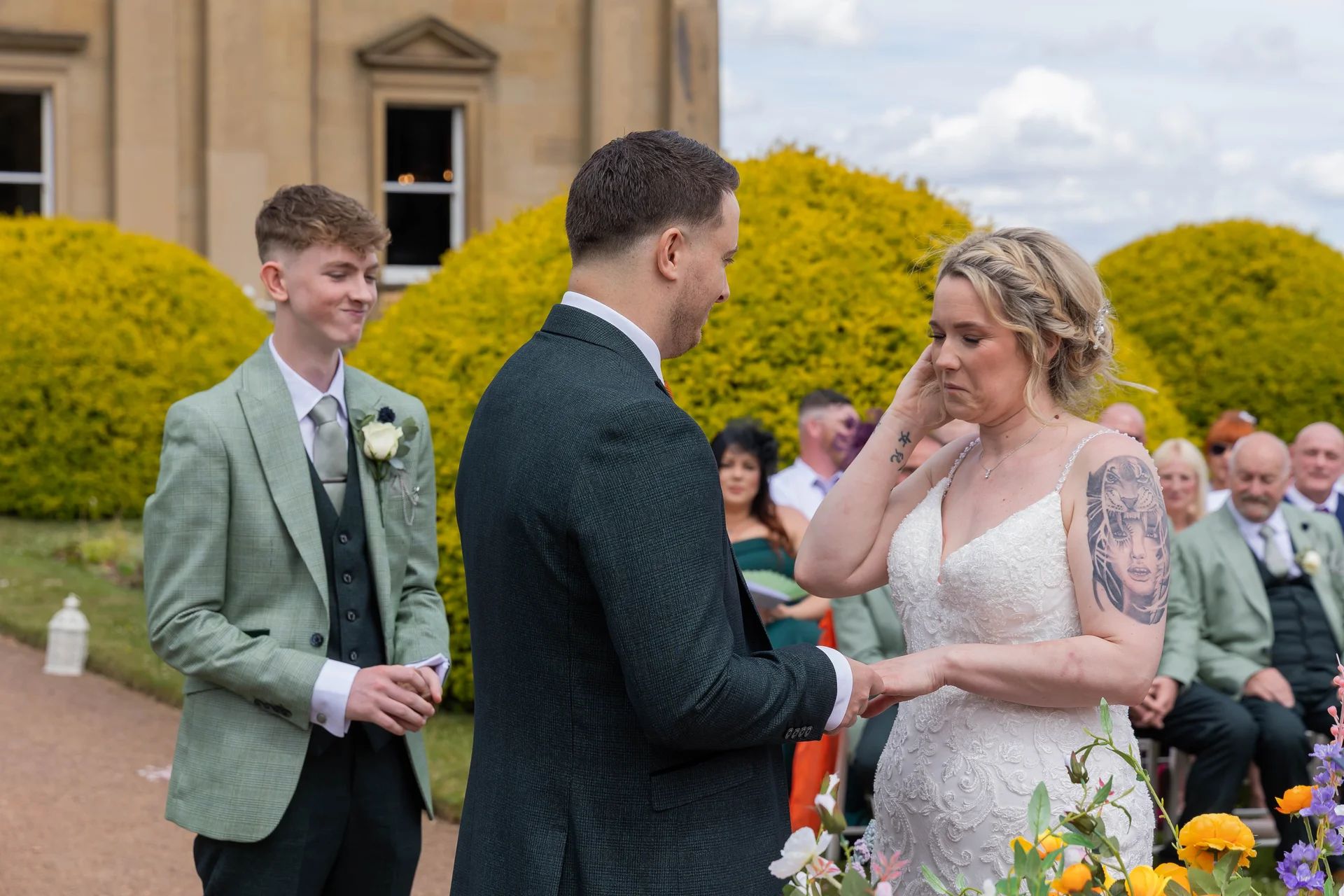 An emotional bride wipes a tear during an outdoor wedding ceremony while holding hands with the groom, watched by a groomsman in a sage green suit.