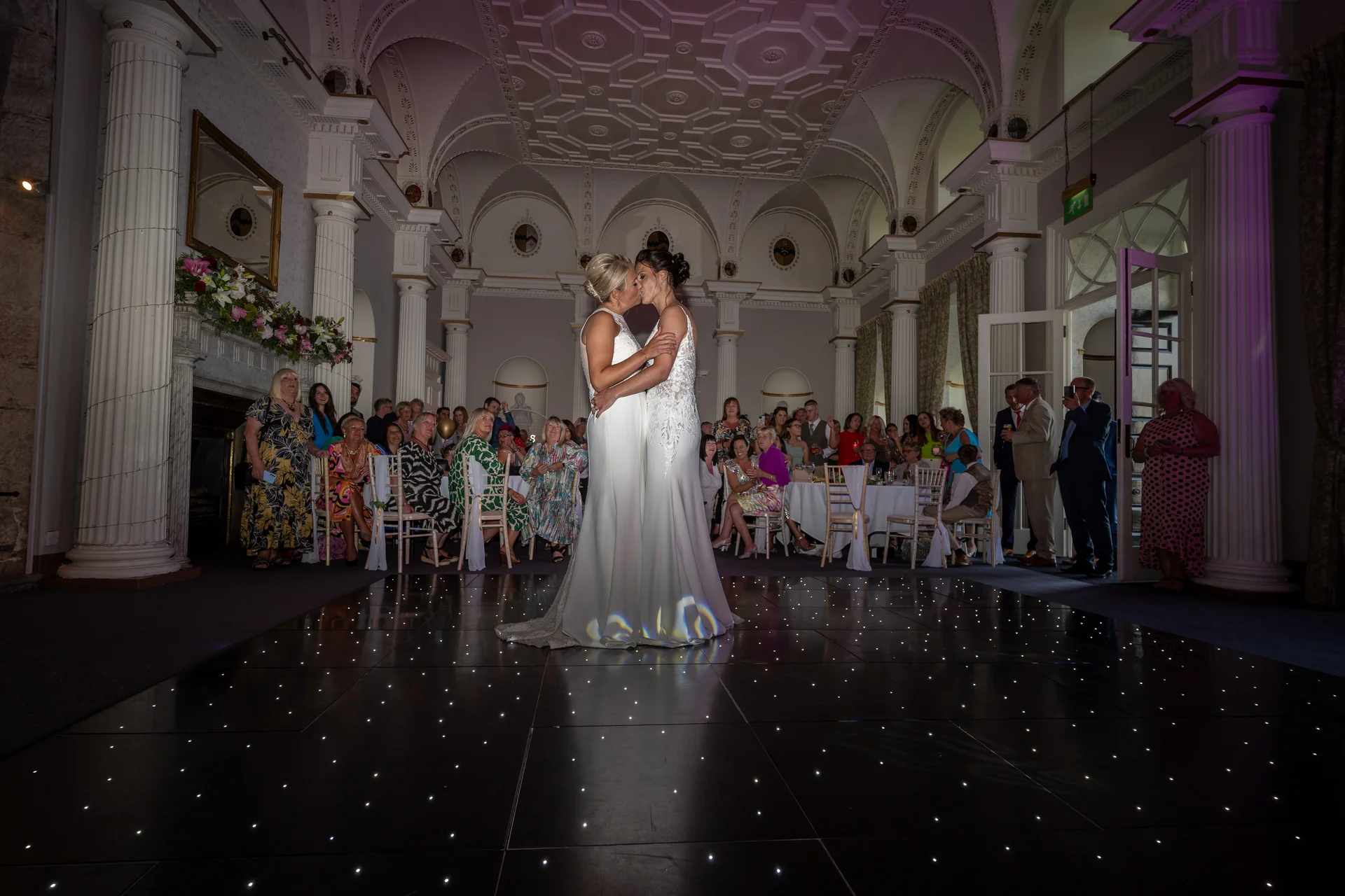 Two brides share their first dance together on a glowing starlit dance floor inside a grand ballroom filled with watching guests.