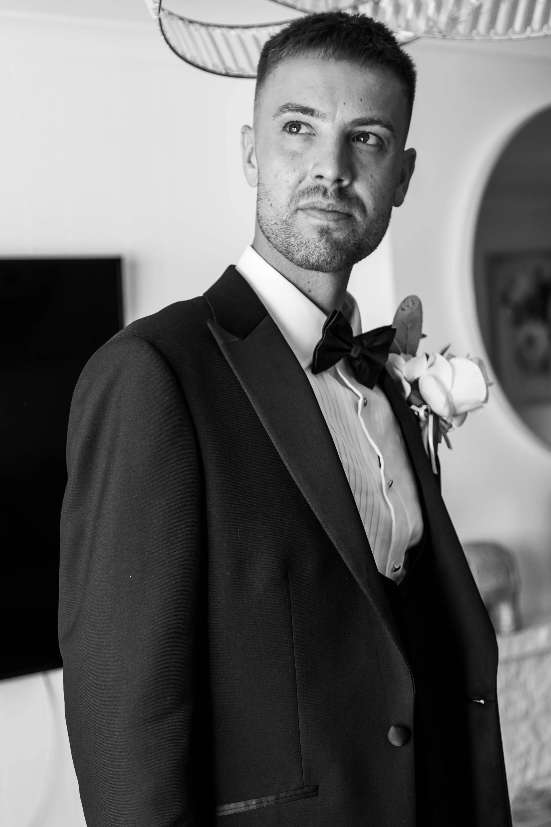 A stylish groom wearing a classic black tuxedo and bow tie looks thoughtfully away from the camera in this elegant black and white portrait.