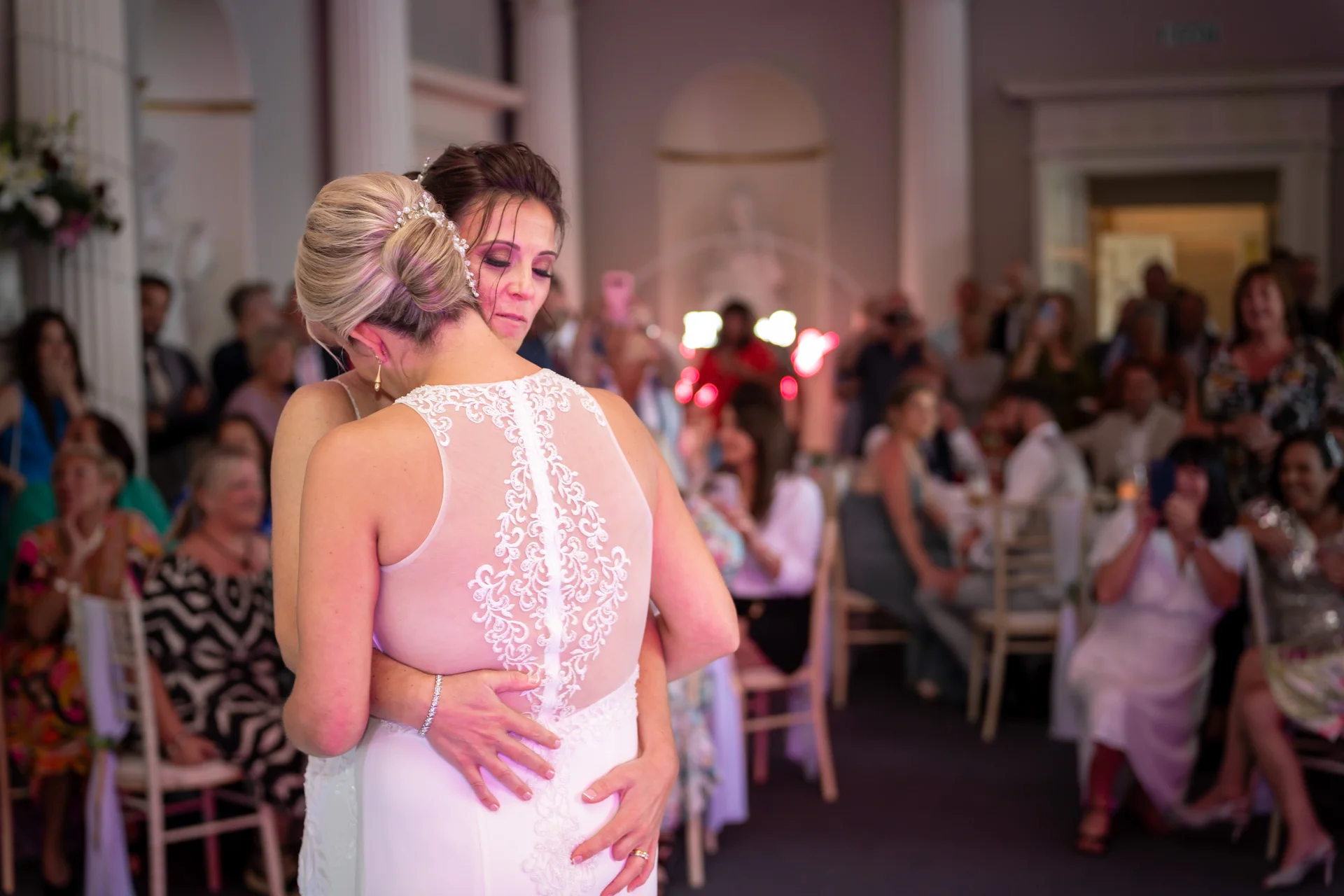 Two brides share a tender embrace during their first dance, surrounded by wedding guests at a reception.