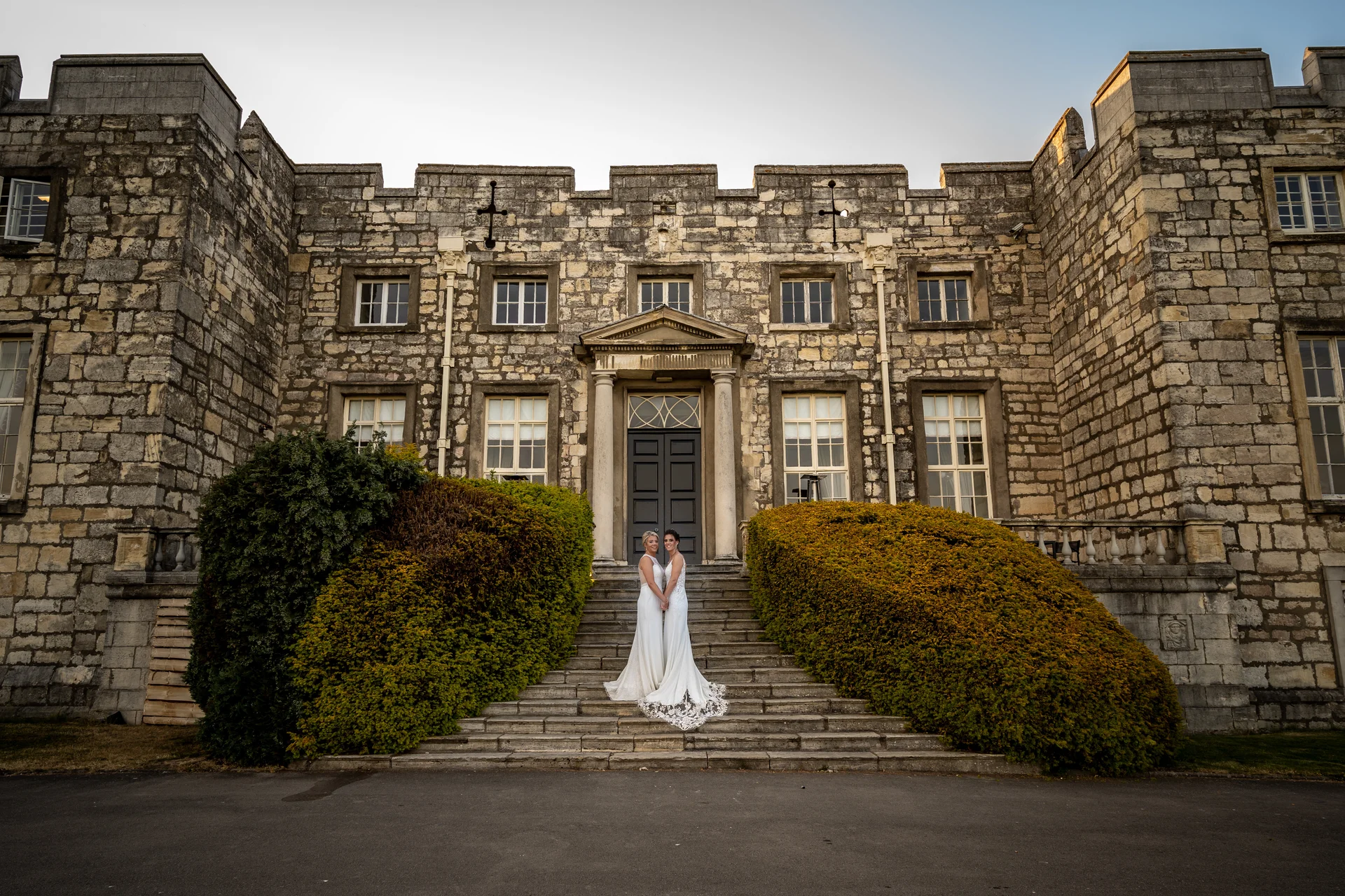 Two brides in elegant white wedding gowns stand hand-in-hand on the grand stone steps of a historic castle venue flanked by manicured shrubbery.