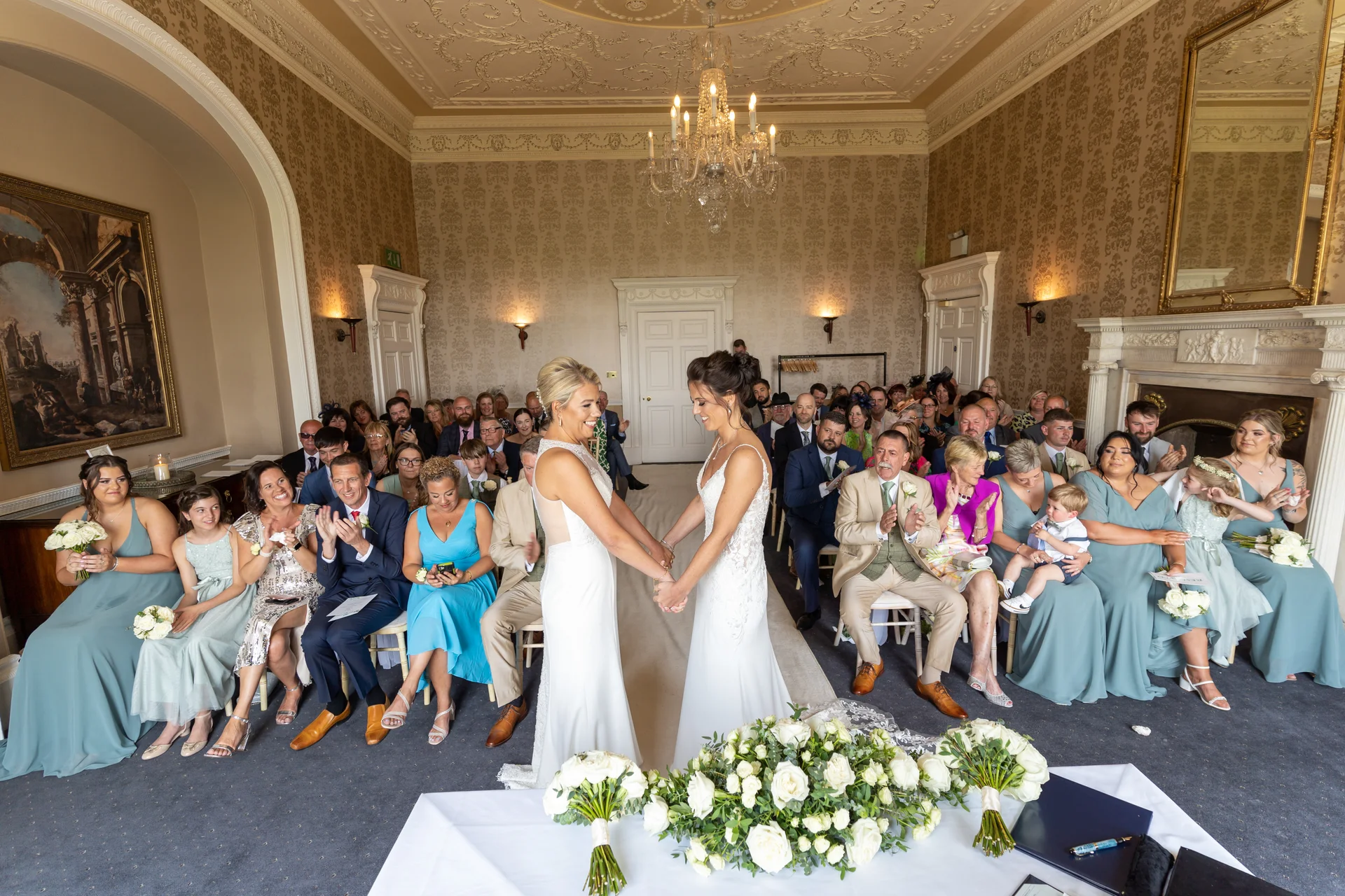 Two brides hold hands and exchange vows during their civil ceremony in a grand stately home drawing room, surrounded by seated guests and a white floral display.