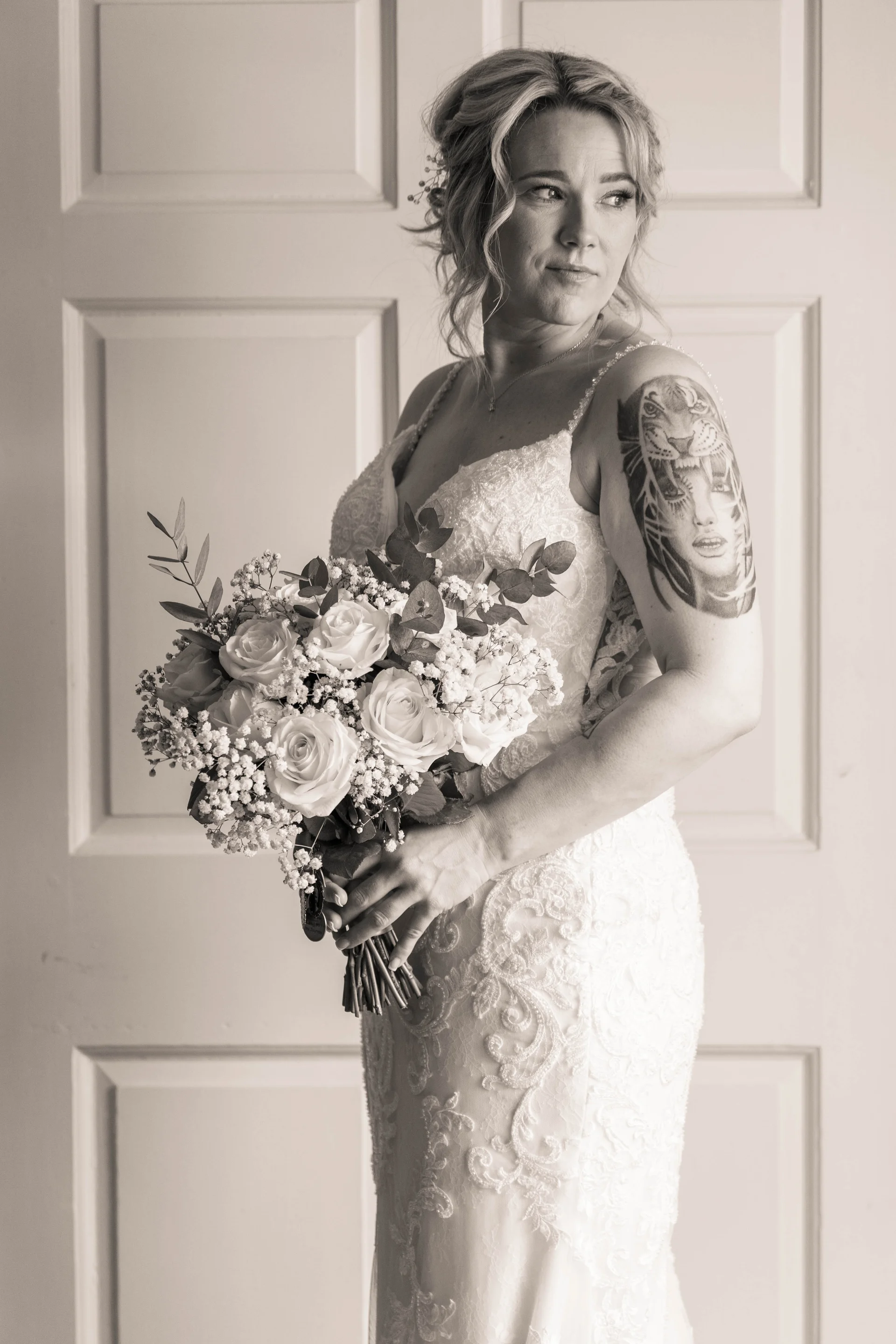 A sepia-toned portrait of a tattooed bride holding a hand-tied rose and baby's breath bouquet while standing before a panelled door.