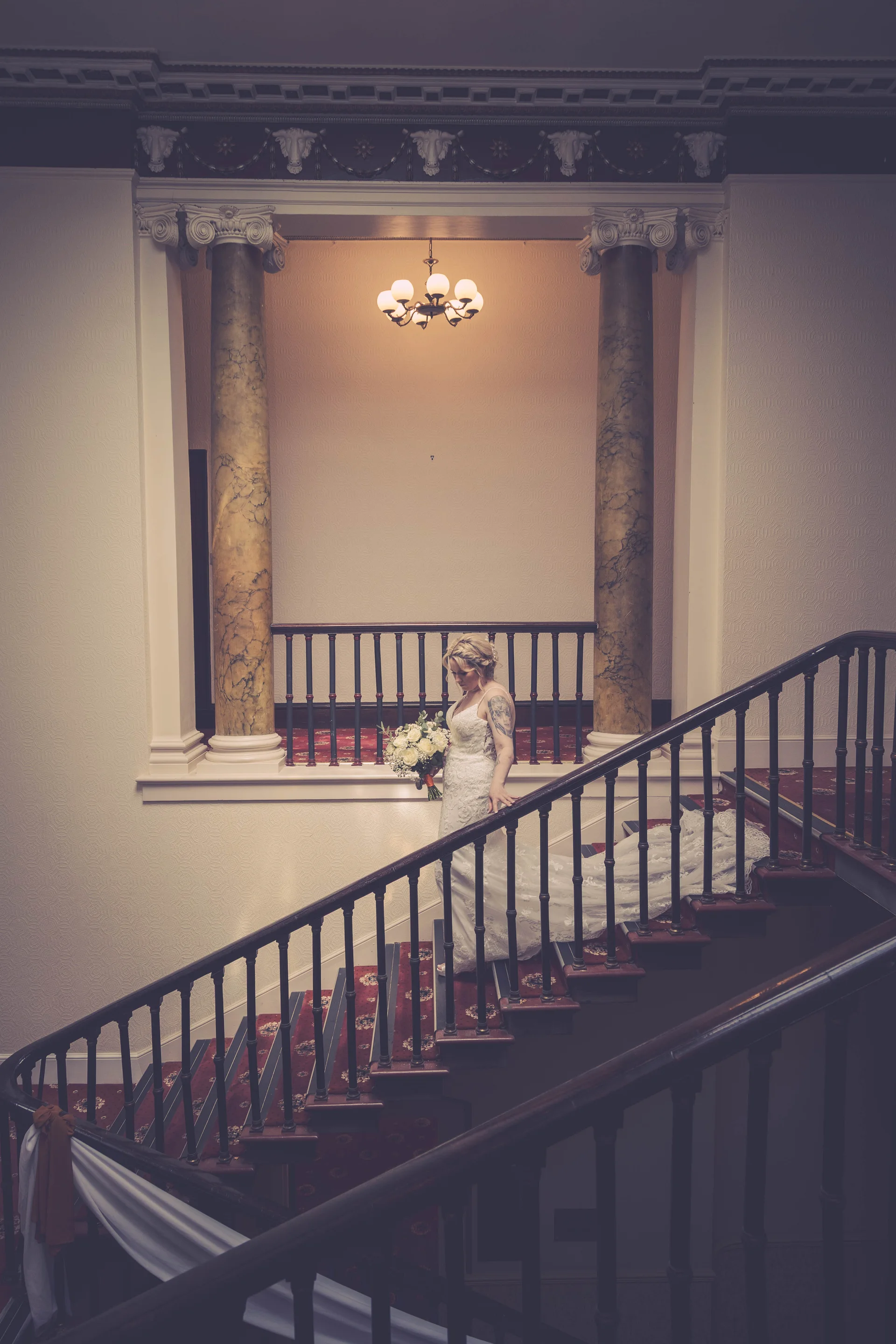 A solo bride wearing a lace strapless a-line gown holding a bouquet stands gracefully on a grand indoor carpeted staircase.