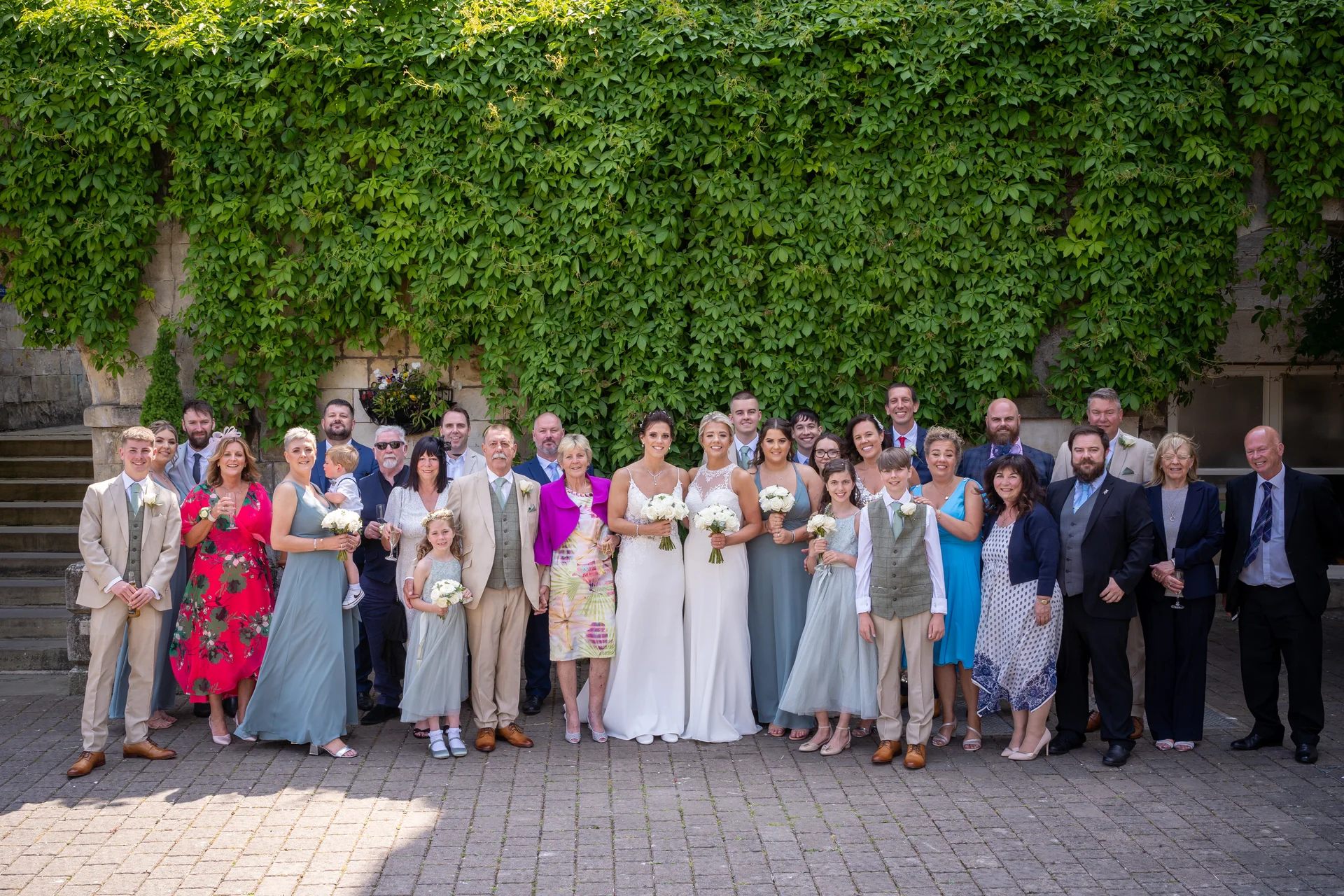 A large wedding party and family group pose together outdoors in front of a tall wall covered in lush green ivy.