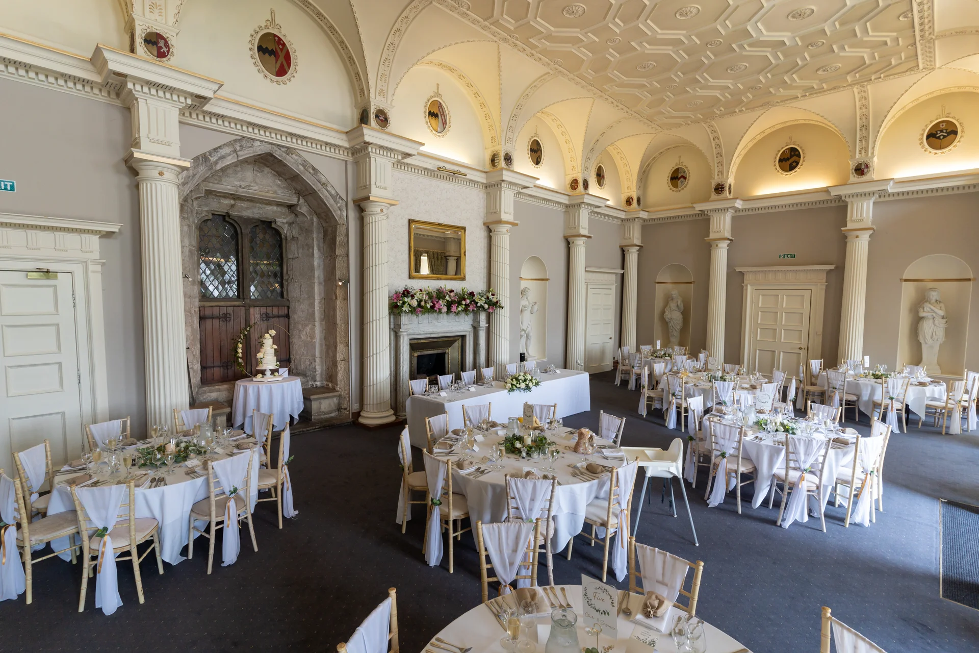 A grand ballroom wedding reception venue features high vaulted ceilings, ornate columns, stone archways, and elegantly dressed round tables with white linens.