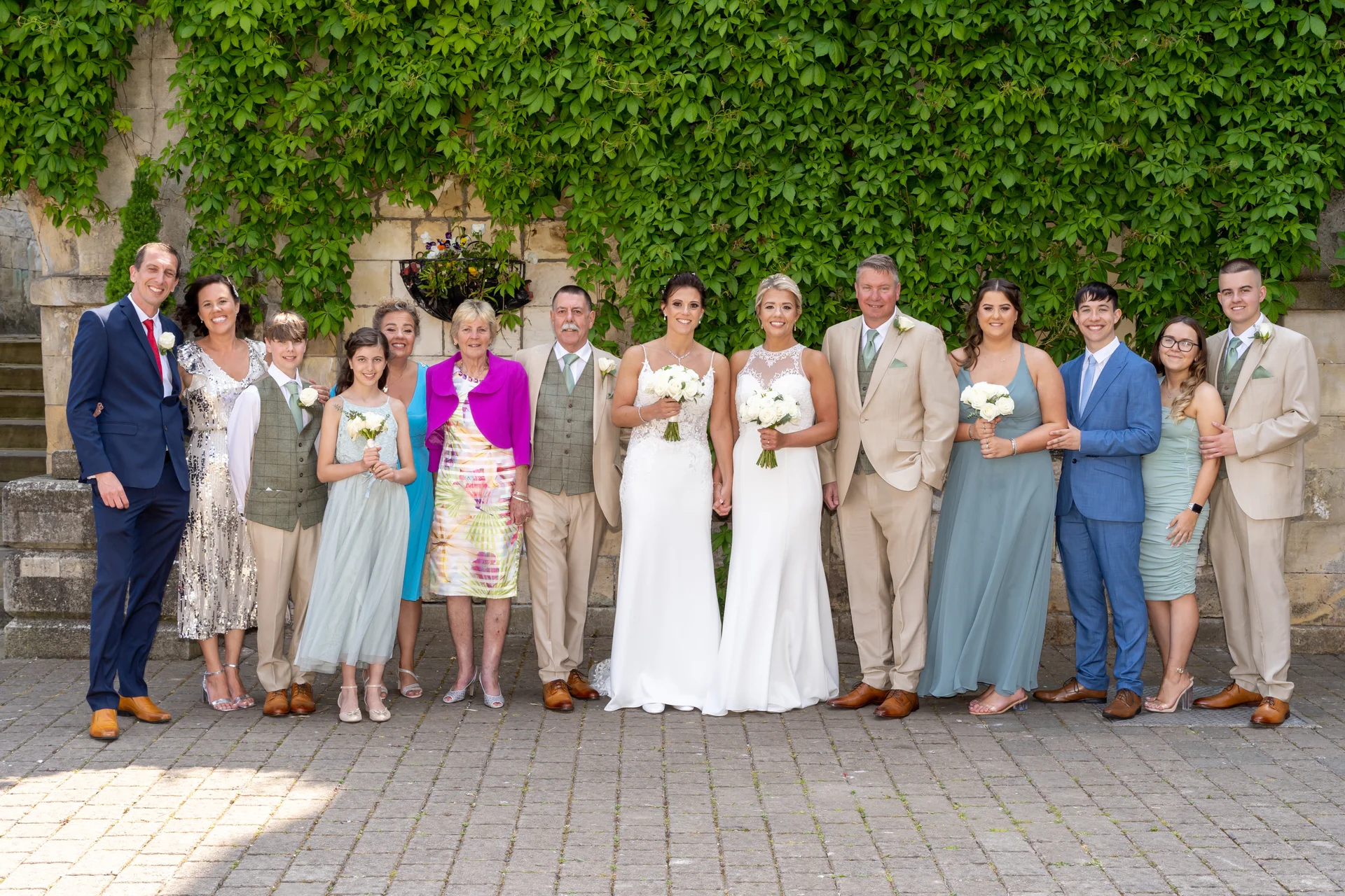 A formal group wedding portrait featuring two brides in white gowns standing centrally surrounded by family members and bridal party members against an ivy-covered stone wall.