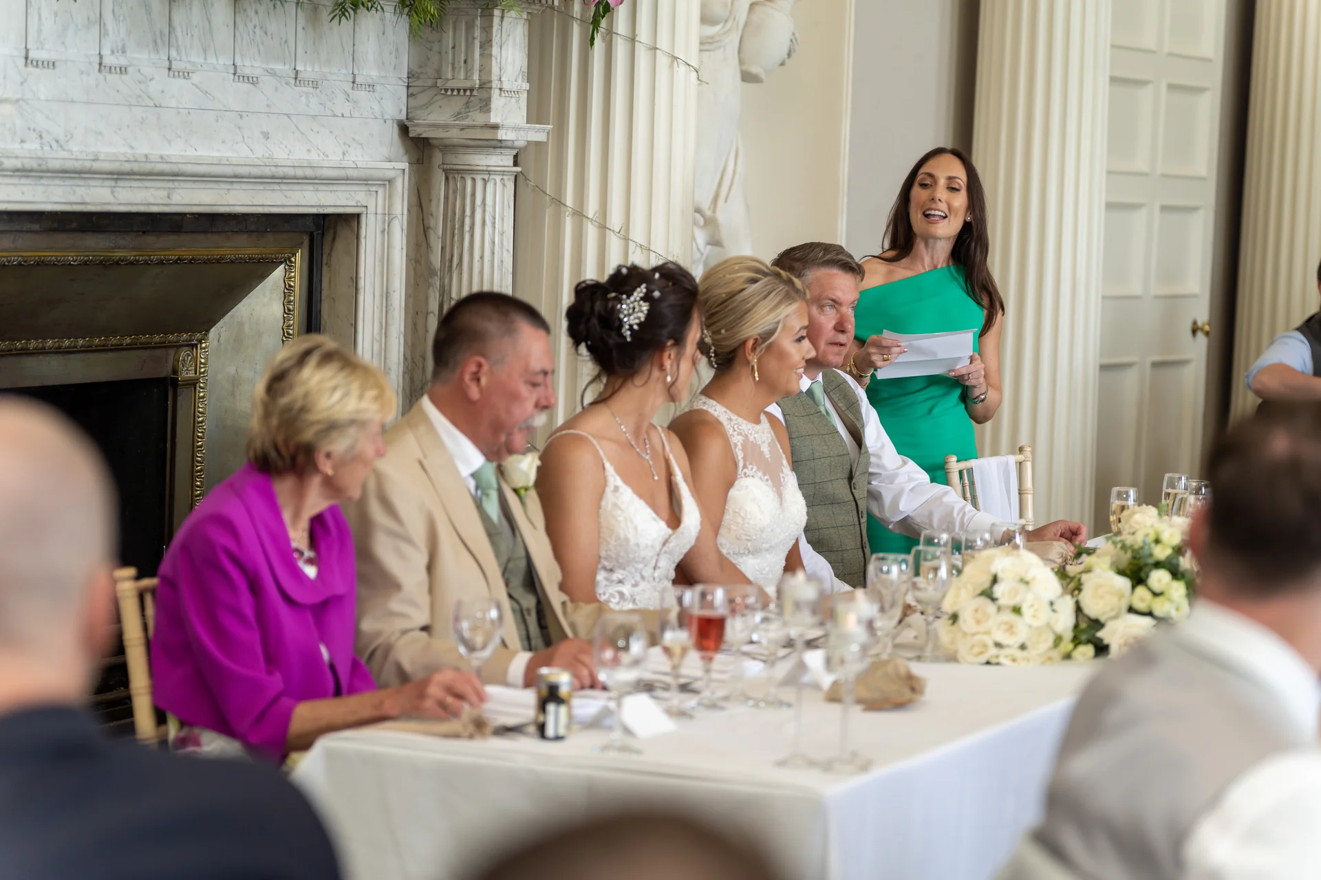 A smiling maid of honour delivers a speech at the top table during a wedding reception in a grand manor house.