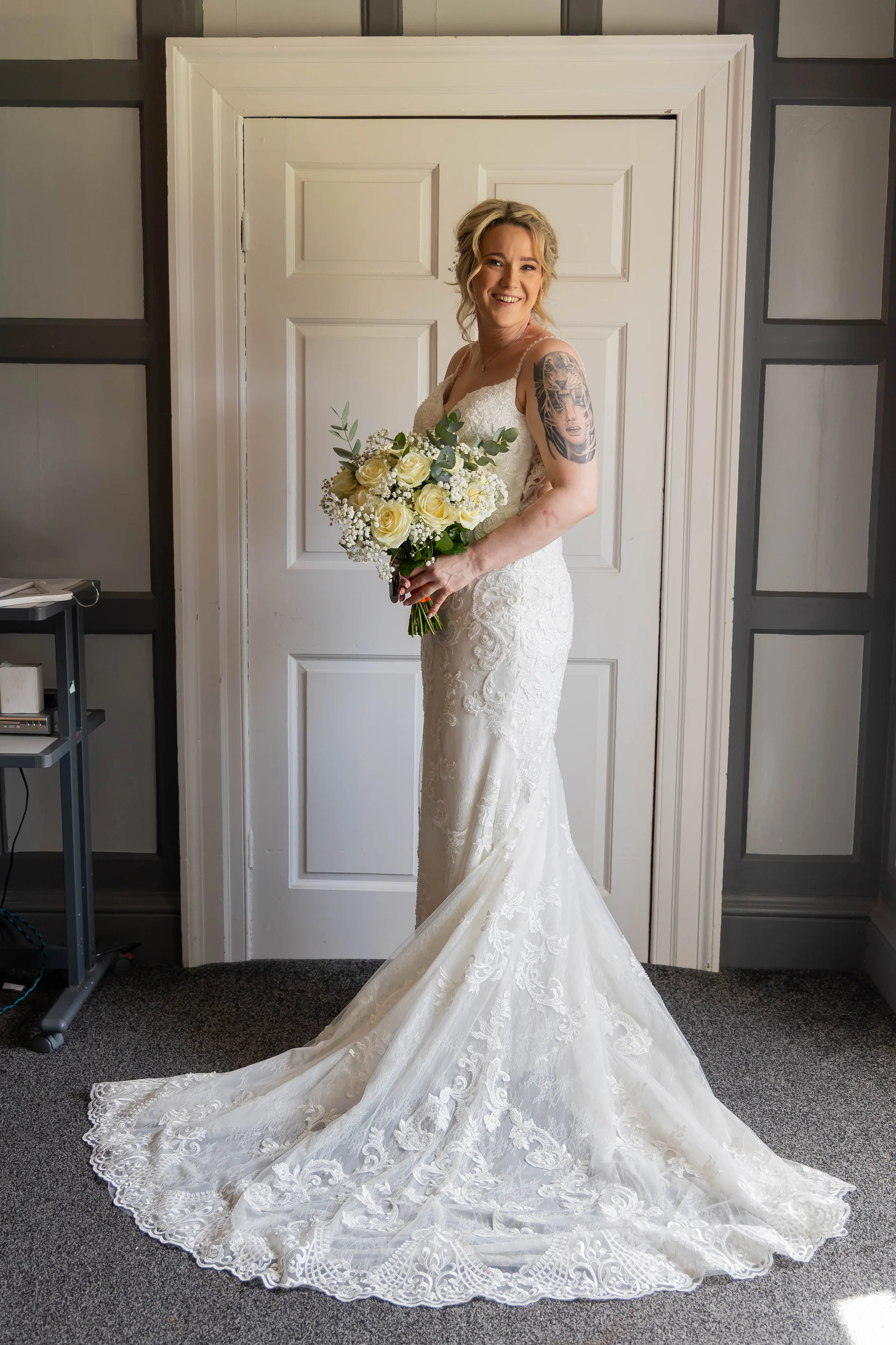 A smiling bride holding a white rose bouquet stands proudly in a lace fit-and-flare gown with a dramatic train inside a panelled room.
