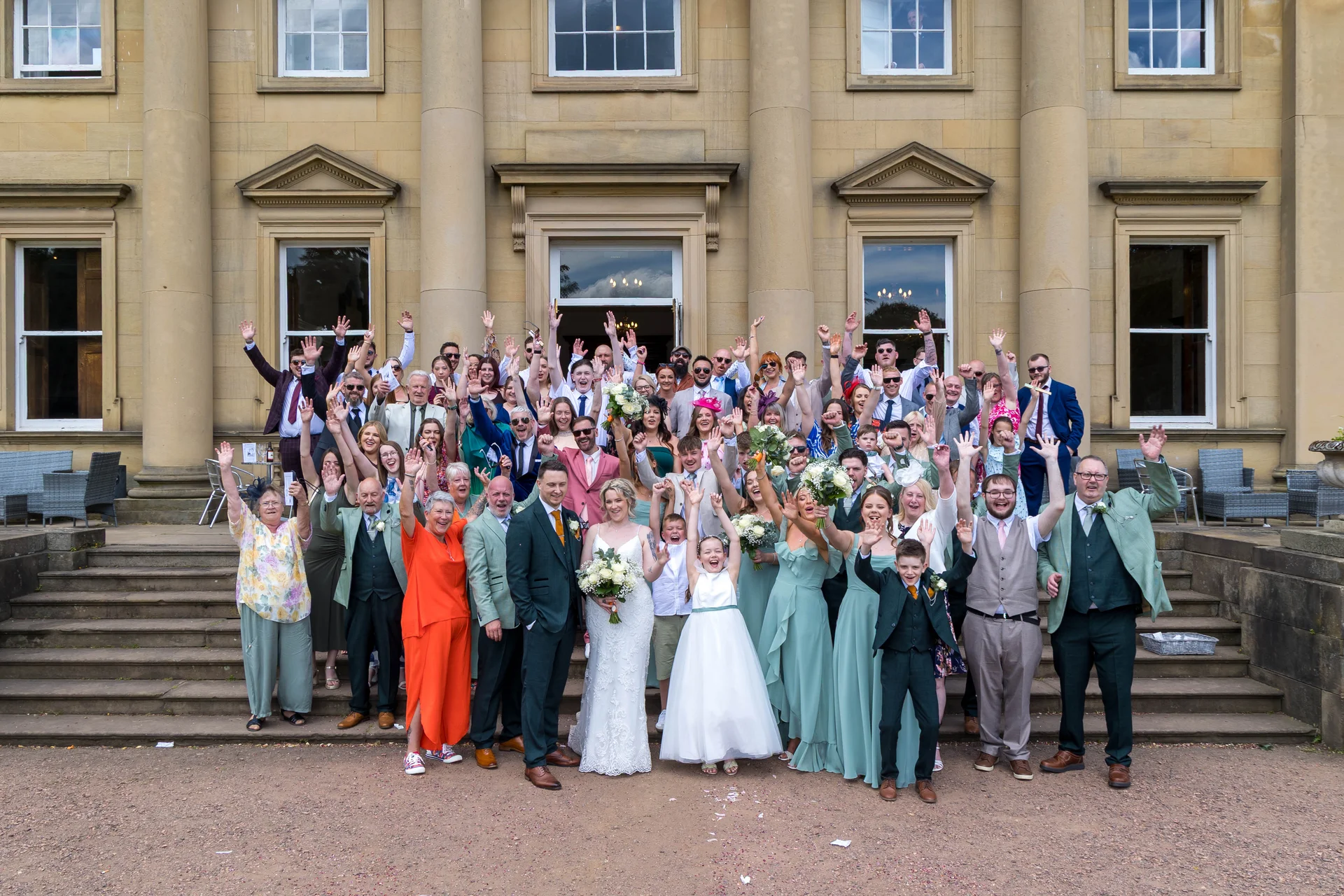 A large wedding party and guests wave enthusiastically while gathered on the grand stone steps outside a stately country house.