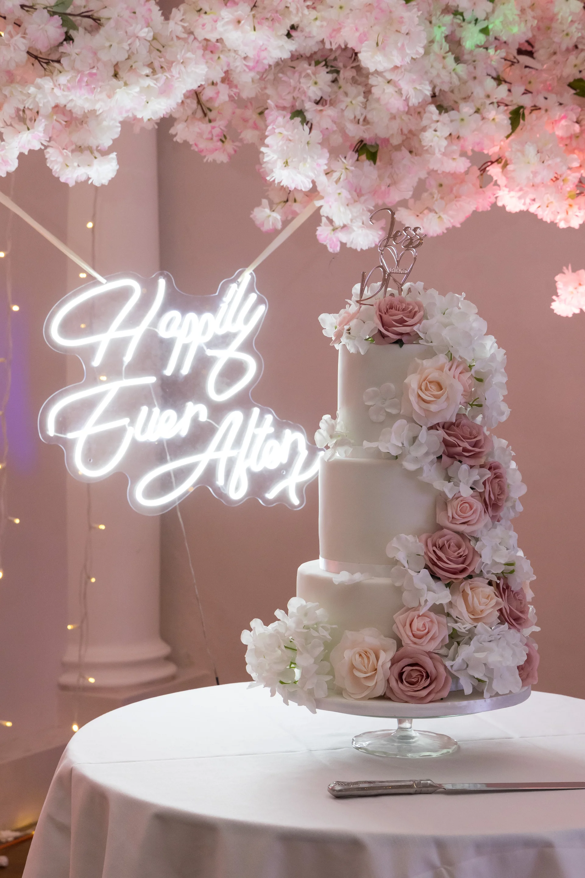 A stunning three-tiered white wedding cake adorned with a cascading arrangement of pink and white roses, positioned beneath a glowing neon sign and a lush cherry blossom floral canopy.