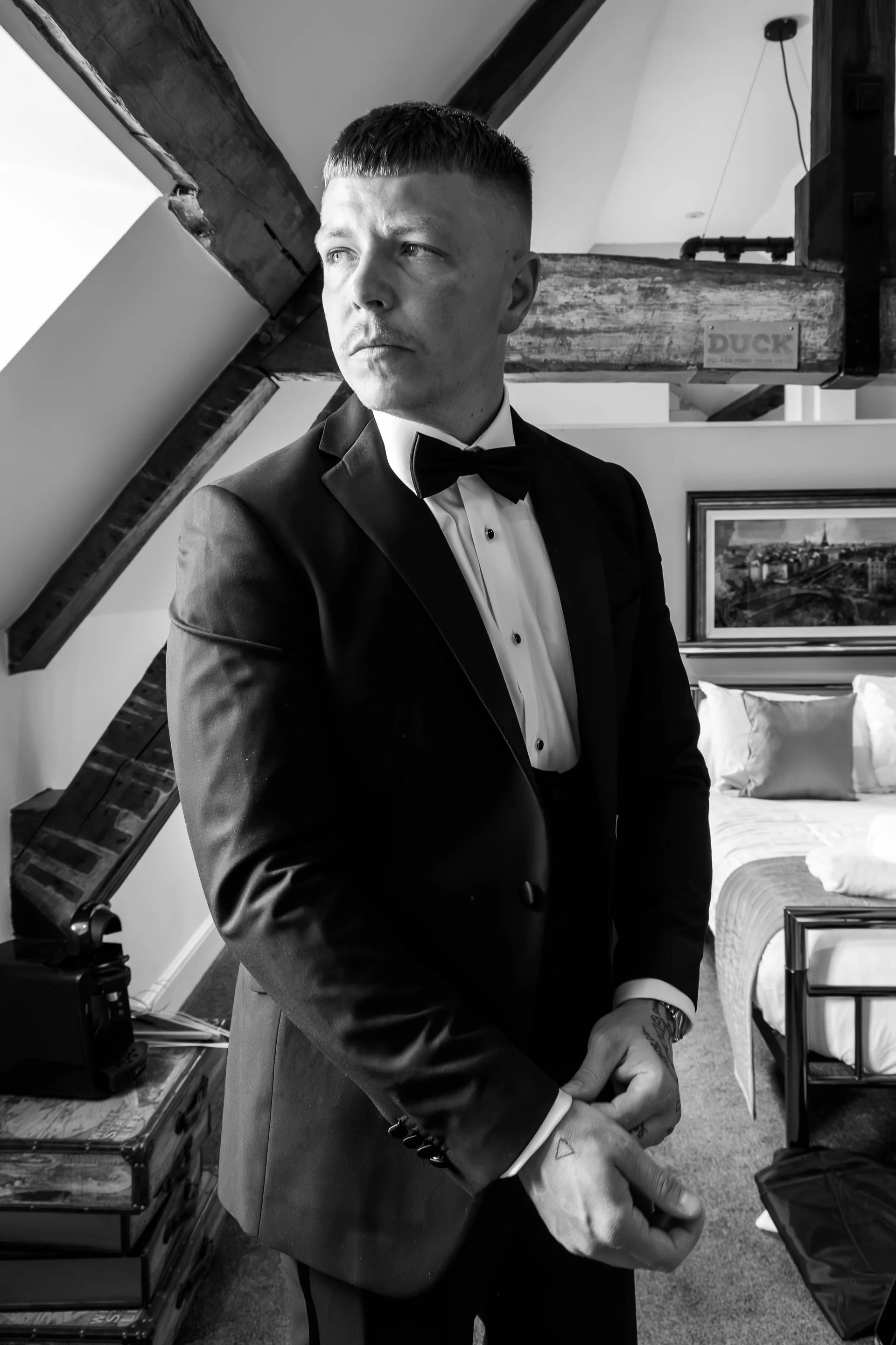 A stylish groom fastens his cufflinks in a light-filled hotel room featuring exposed wooden rafters while getting ready for his wedding day.