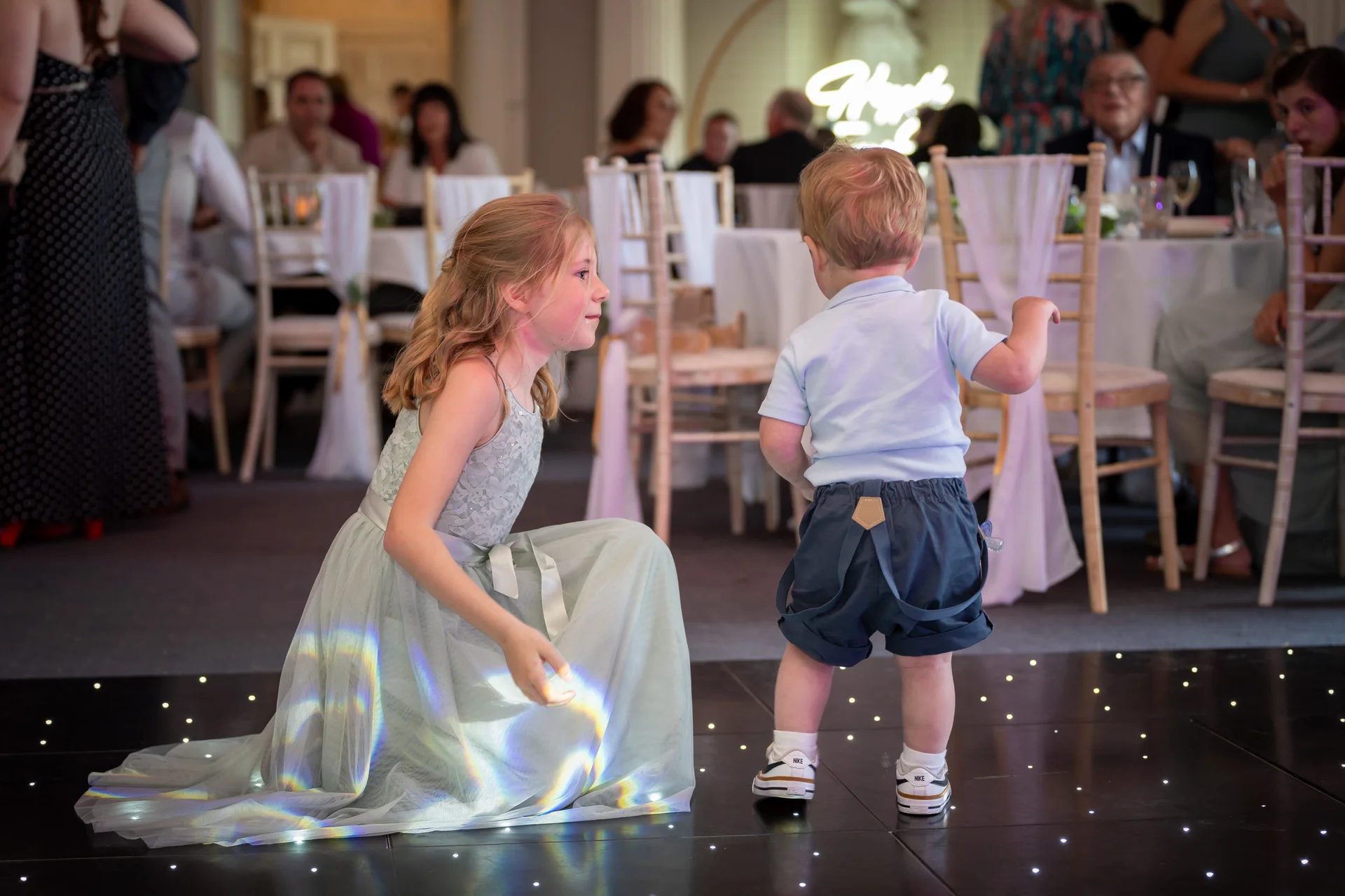 A young flower girl kneeling on an illuminated dance floor while interacting with a page boy at a wedding reception.