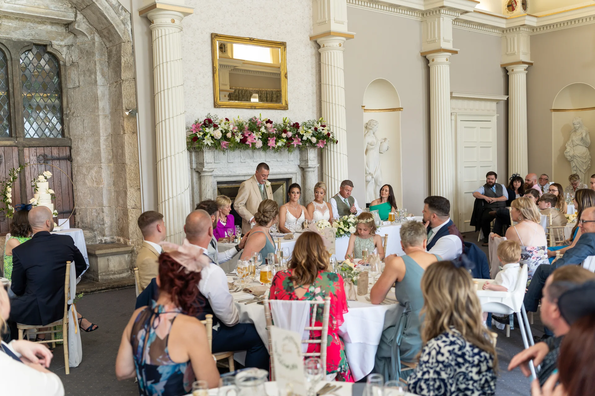 A wedding speech is delivered at the top table in a grand ballroom featuring classical pillars and ornate fireplaces, with seated guests listening attentively.