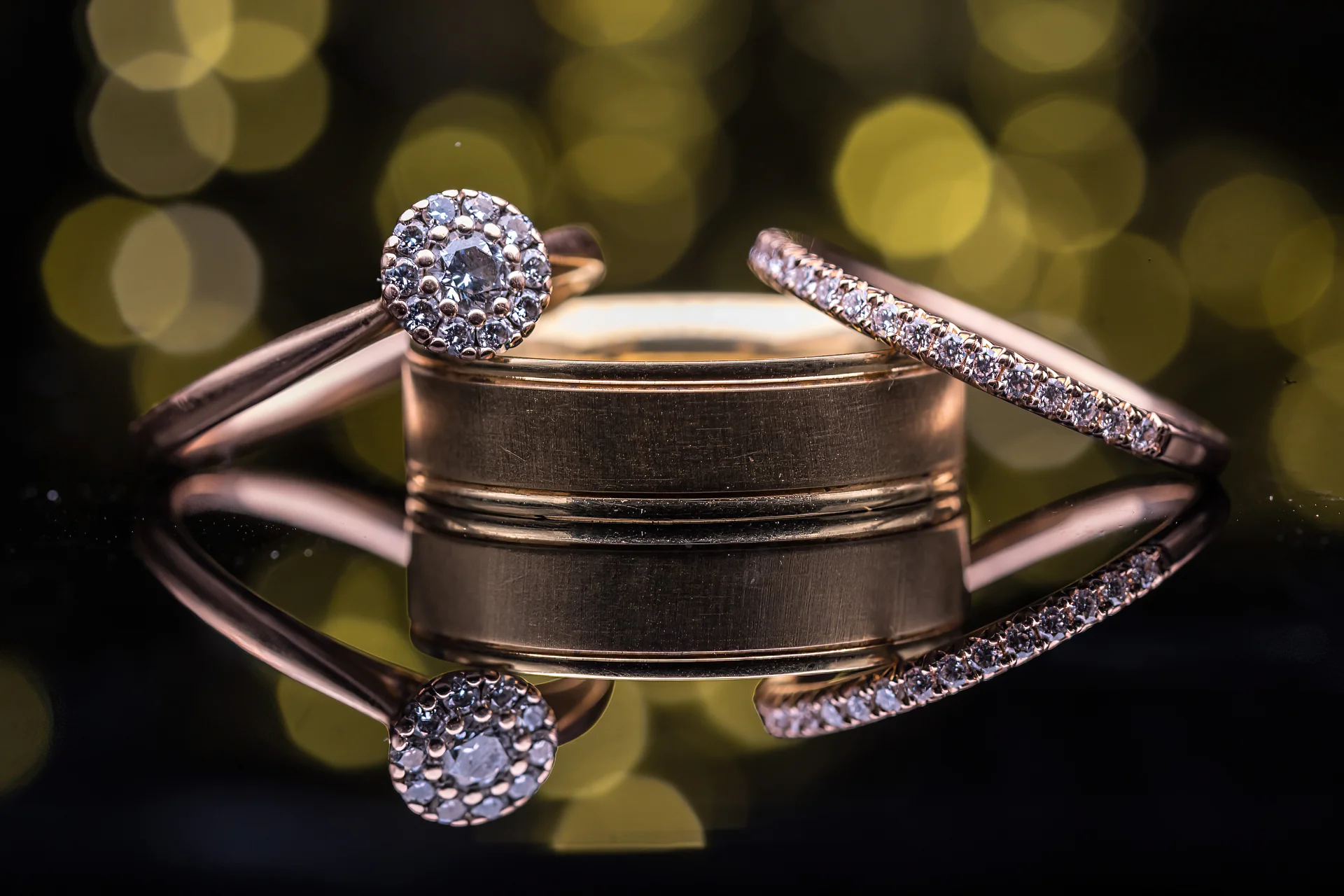 A macro detail shot of sparkling wedding rings and a diamond engagement ring displayed on a reflective surface against a warm bokeh background.