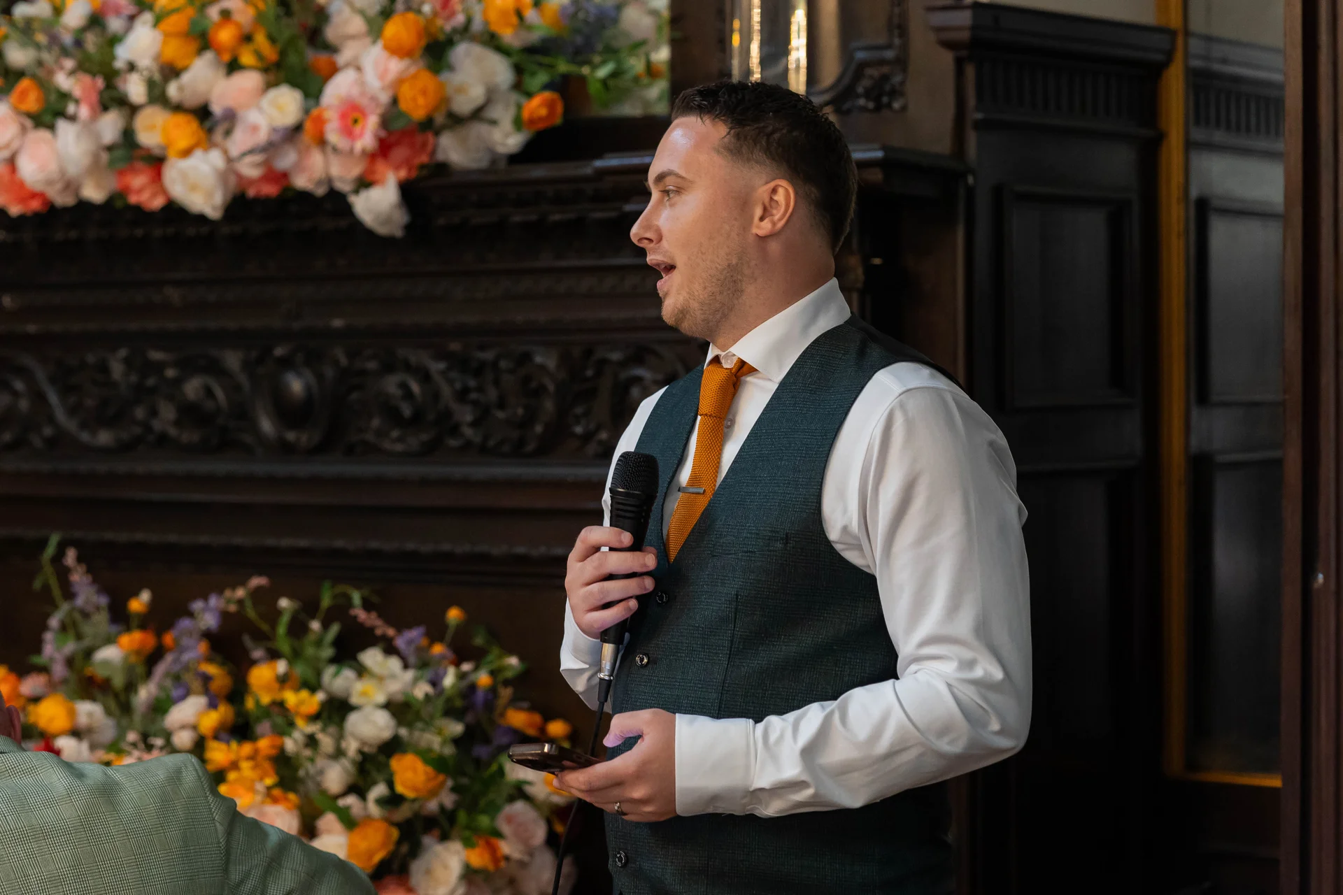 A groom wearing a waistcoat and orange tie delivers a speech into a microphone in front of a large floral fireplace arrangement.