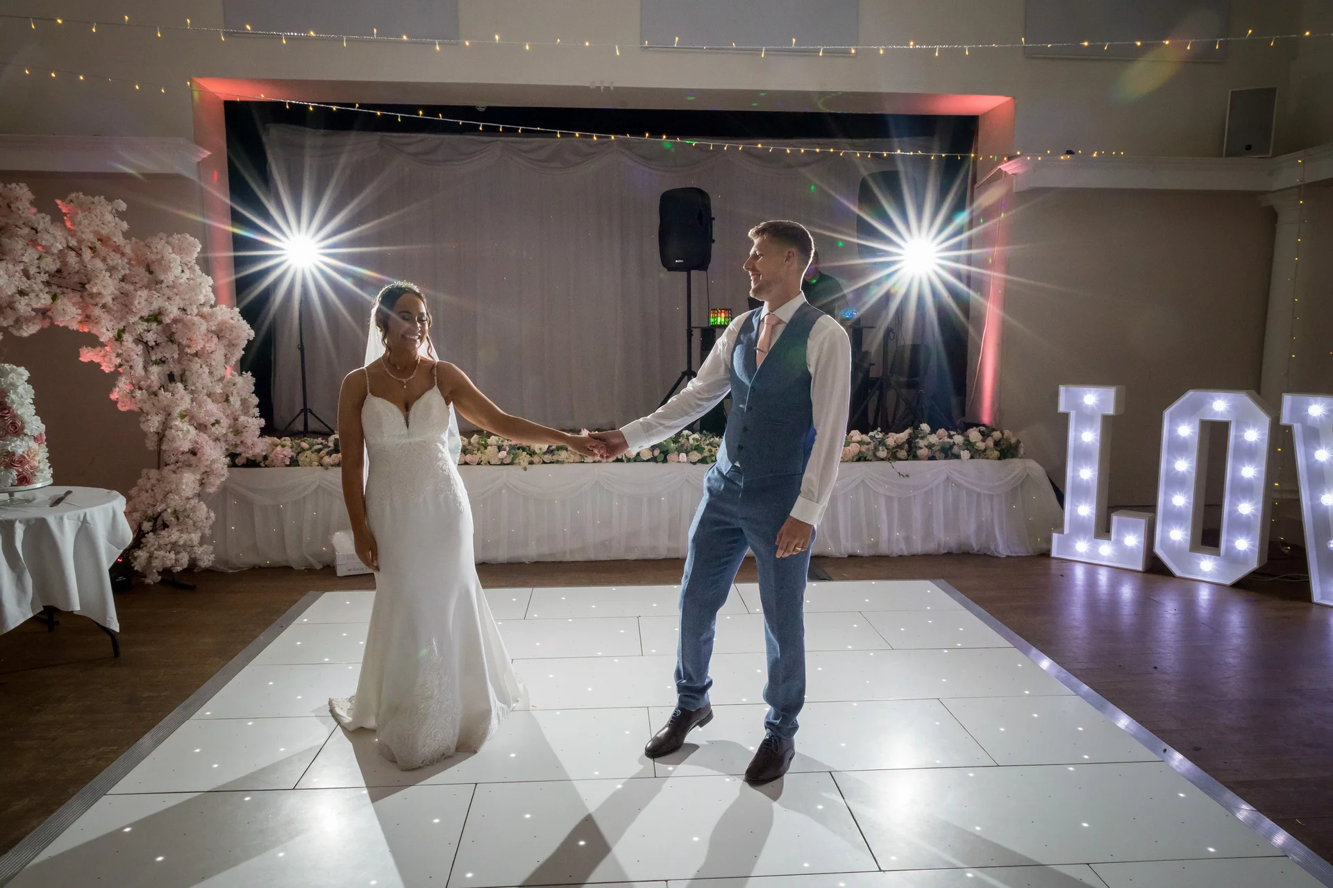 A newlywed couple shares their first dance on a glowing white dance floor under starburst stage lights inside a village hall.