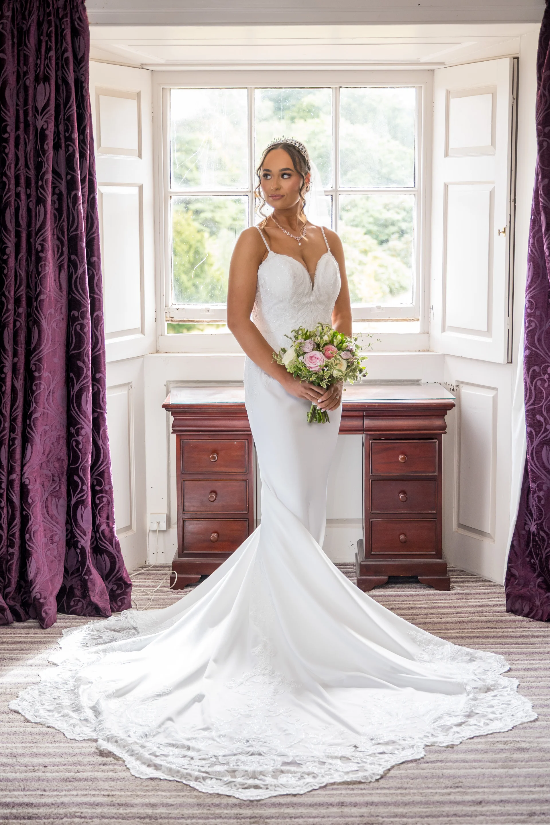A stunning bride poses gracefully by a window in a fitted mermaid wedding gown with a dramatic lace-edged train, holding a delicate hand-tied bouquet.