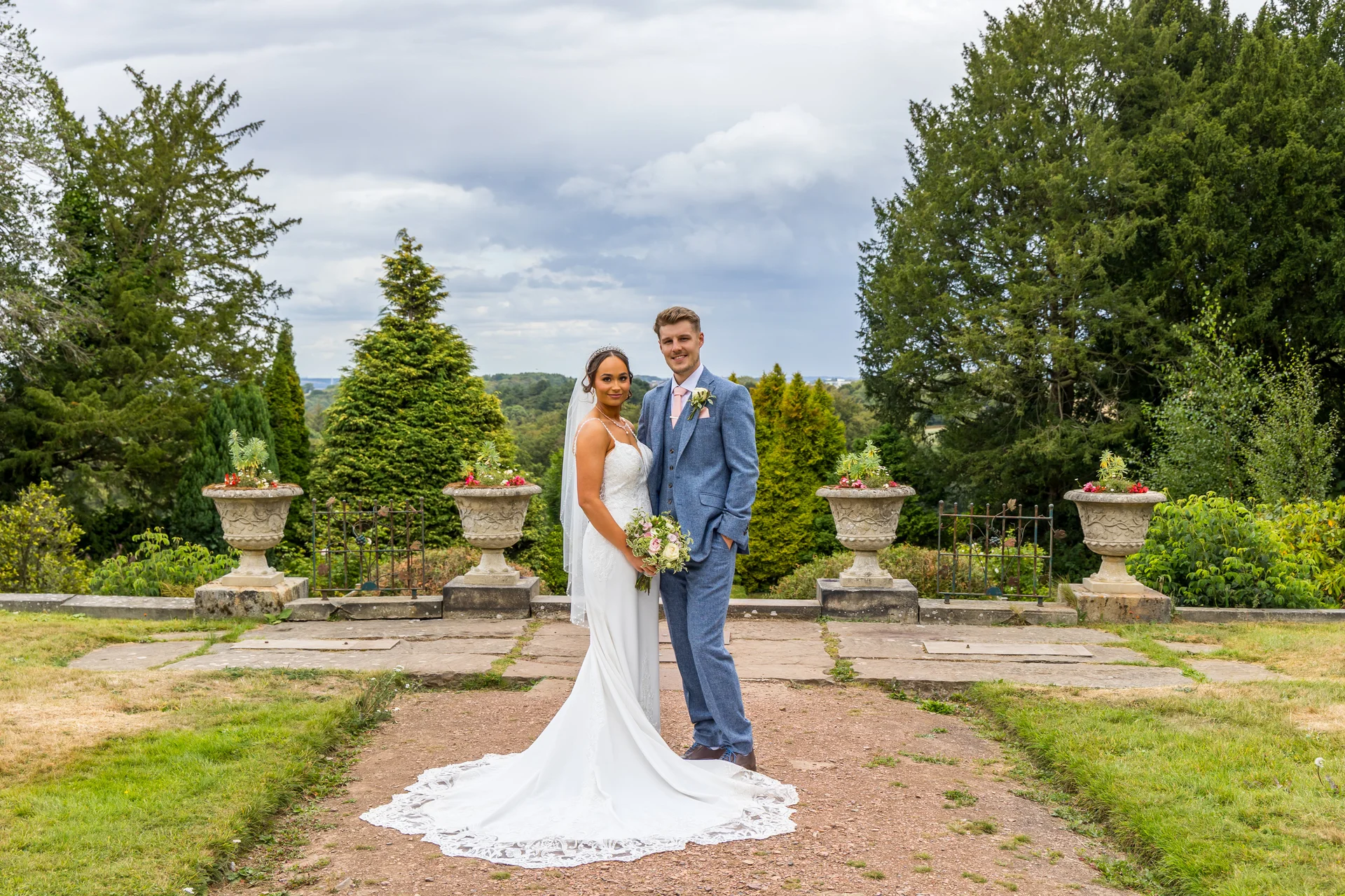 Newlywed bride and groom pose together on a stone terrace in a manicured garden with urns and a dramatic cloudy sky overhead.