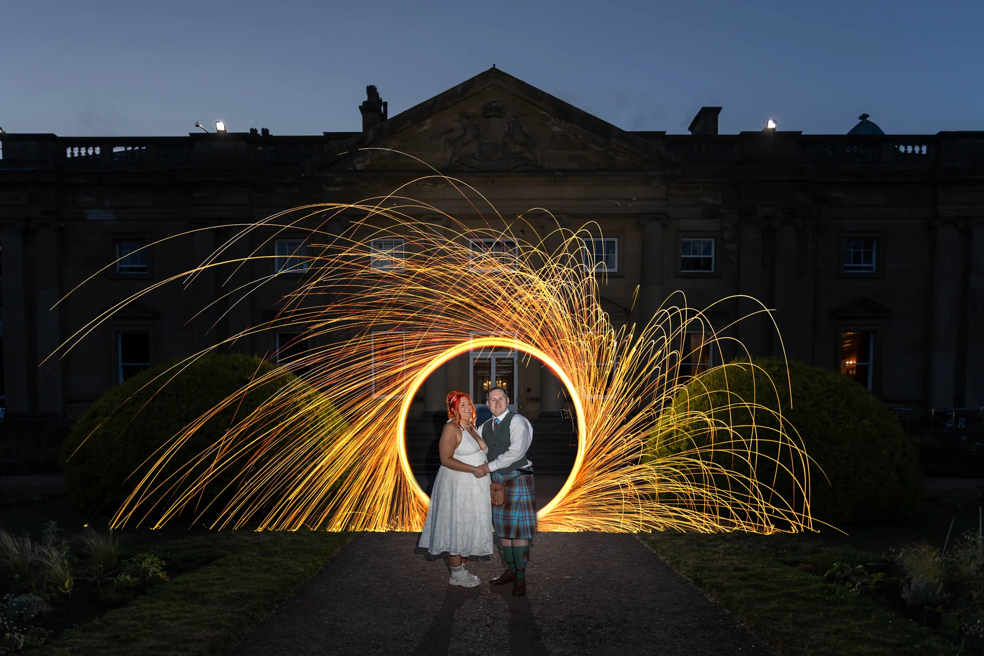 A bride and groom stand together on a path at night in front of a grand country house, framed by a spectacular spinning circle of sparkler light painting.