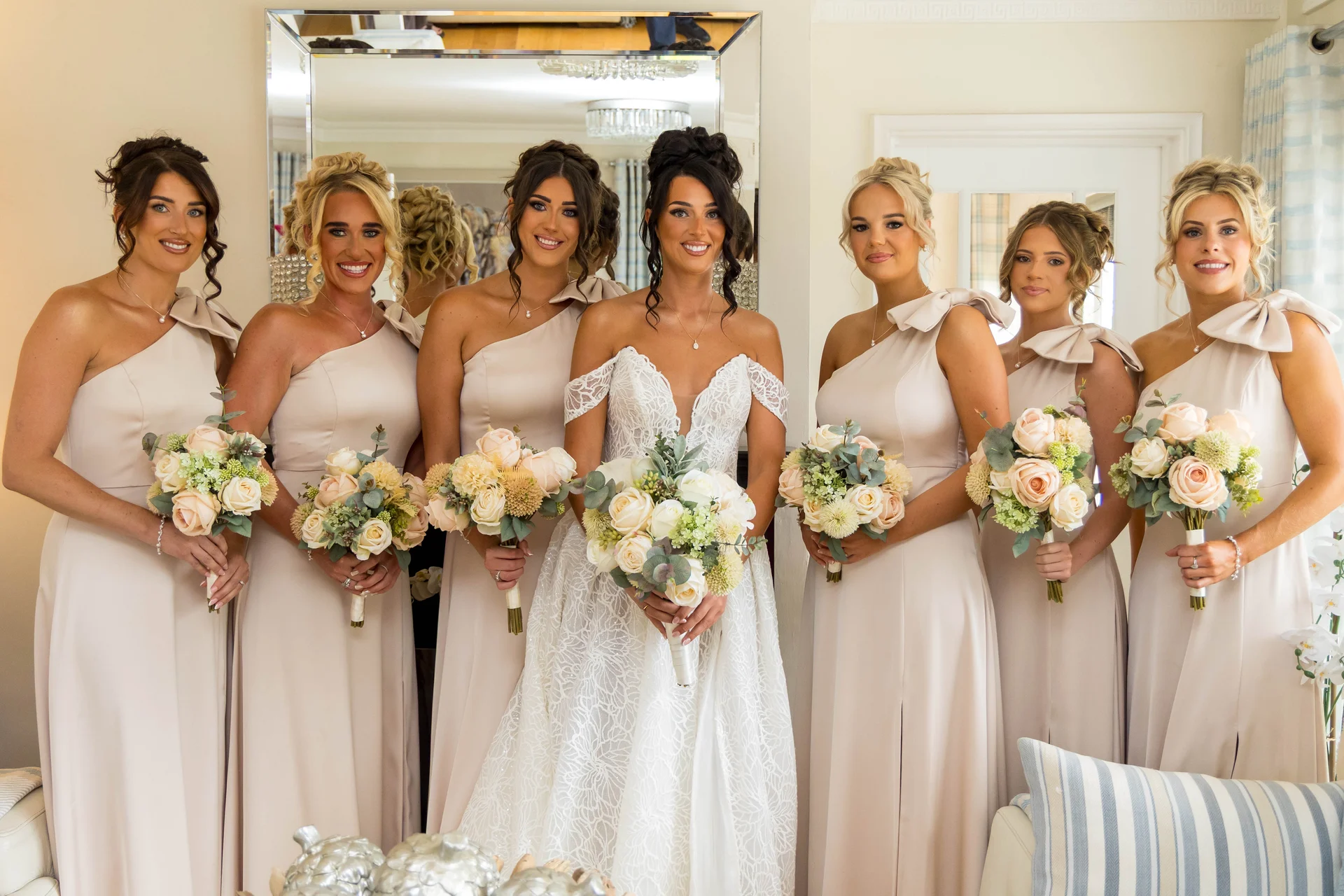 The radiant bride stands proudly at the centre holding a lush cream rose bouquet, surrounded by six smiling bridesmaids in matching satin one shoulder champagne gowns.