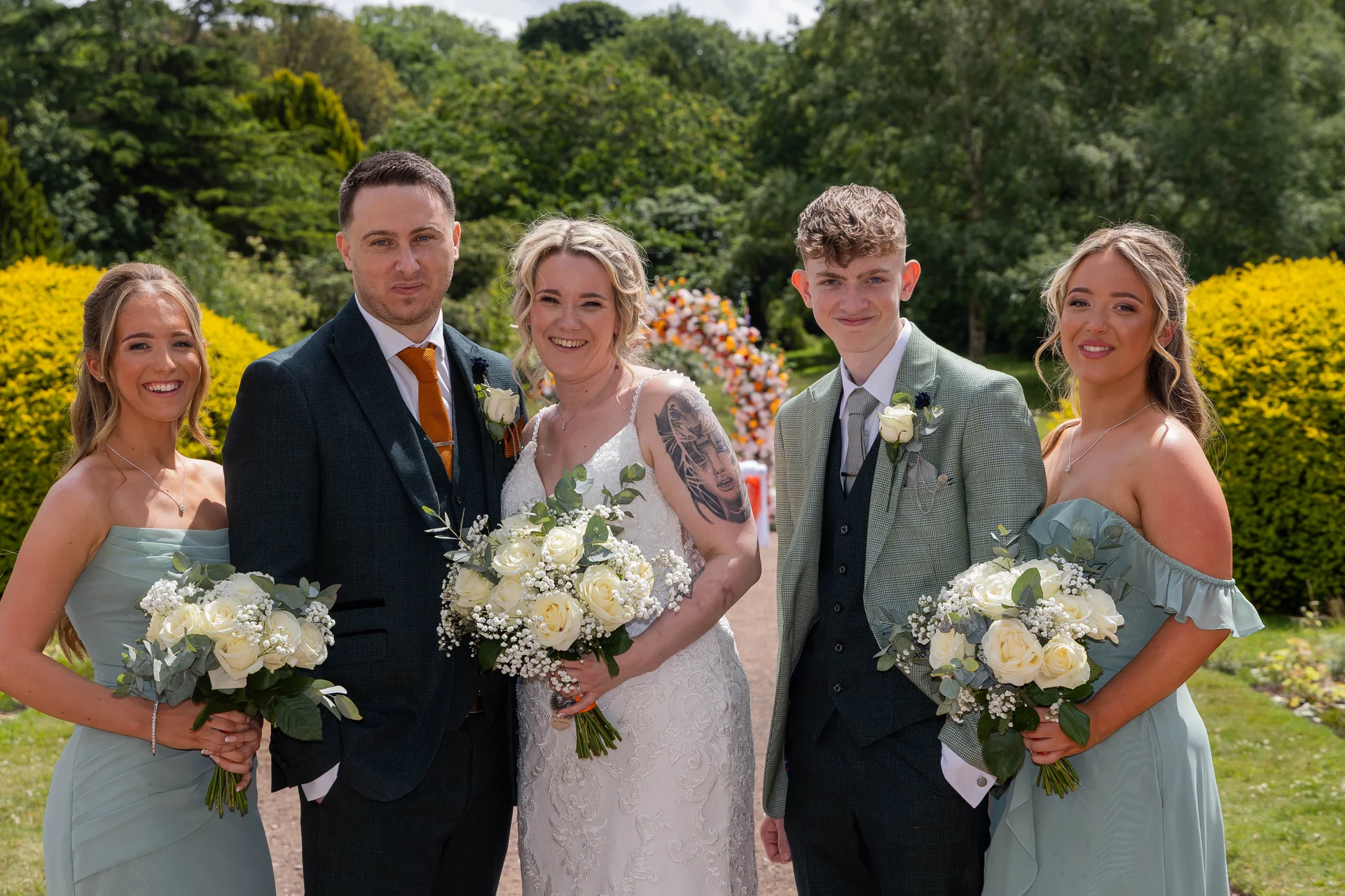 A smiling bride holding a white rose bouquet poses alongside her groom, groomsman and bridesmaids in matching sage green dresses within a sunny garden setting.